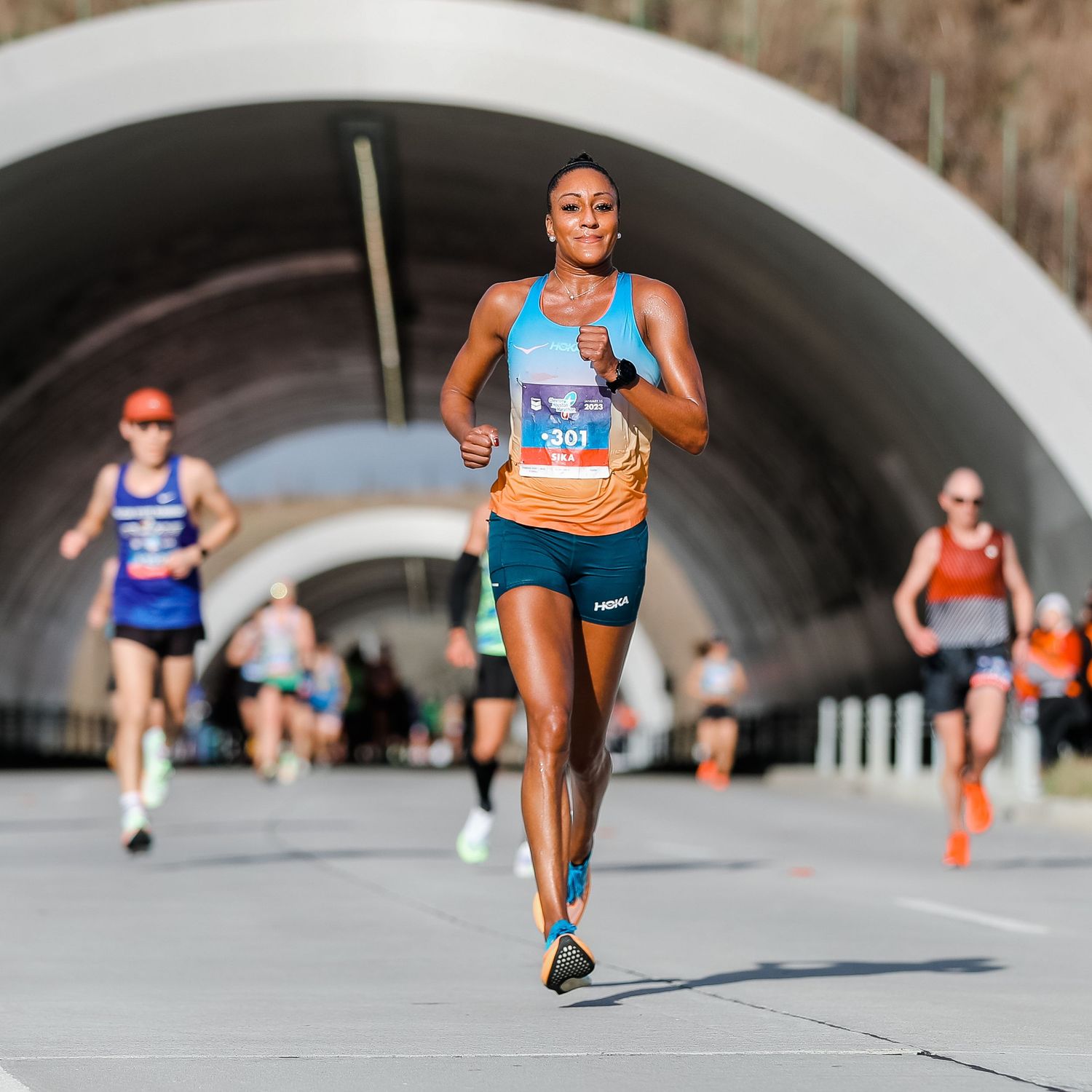 Female runner wearing bib 391 leads a road race through a tunnel arch in athletic gear.