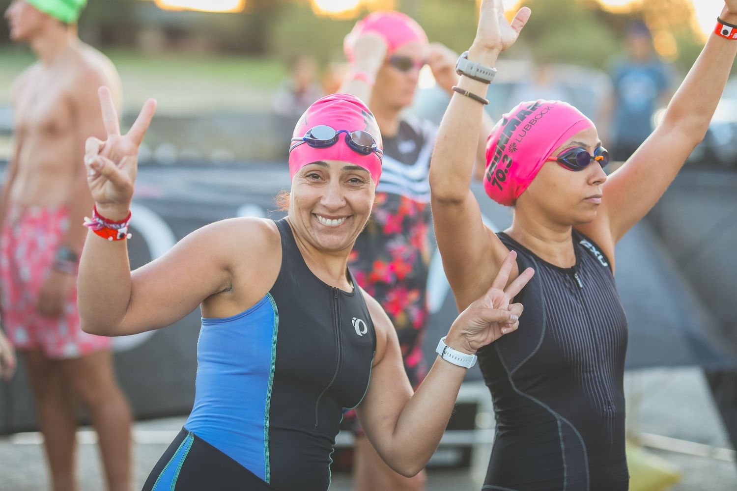 Two women in wetsuits and pink swim caps flash peace signs at triathlon swim start.