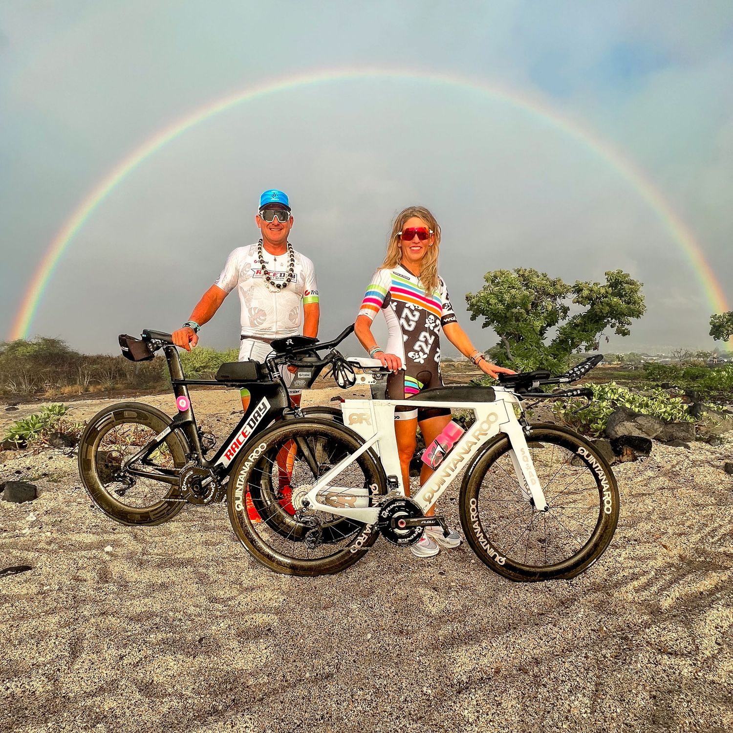 Two cyclists posing with triathlon bikes under a vibrant rainbow on a rocky landscape.
