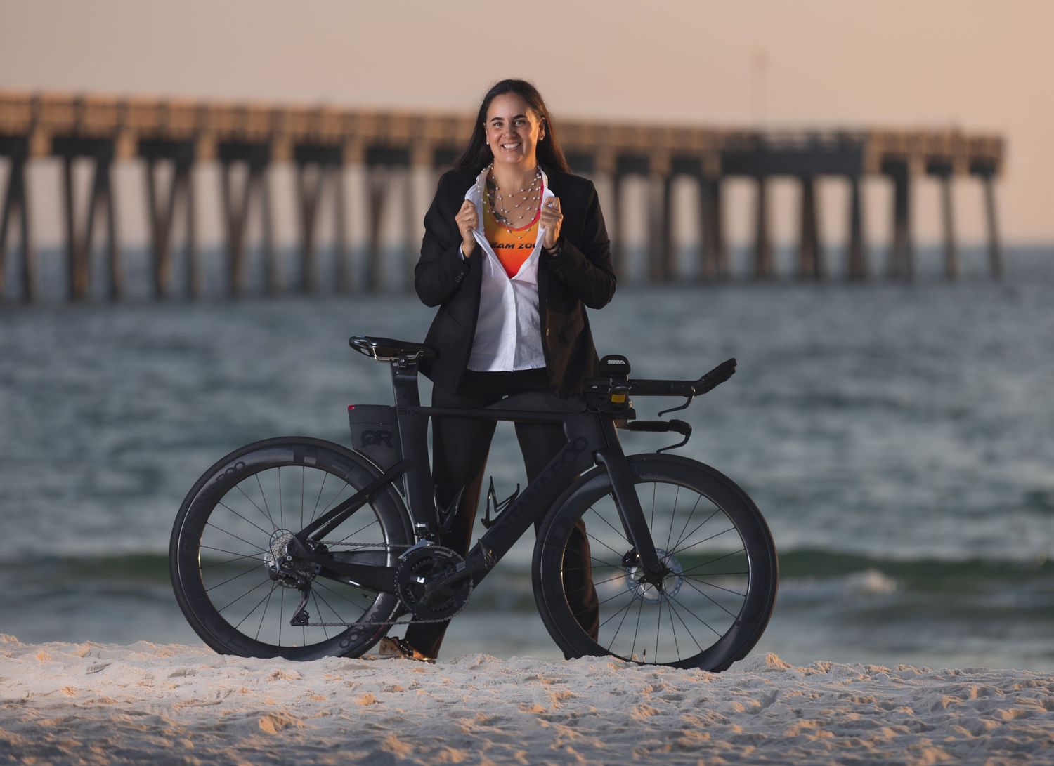 Female athlete posing with triathlon bike on beach near pier at sunset.