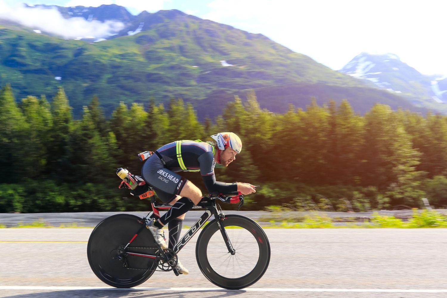 Cyclist in aerodynamic position races on road bike past lush mountain scenery.