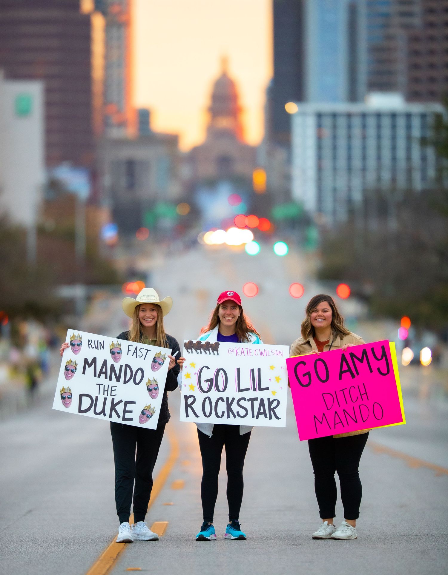Three women hold cheering signs on a city street at sunset during a marathon race event.