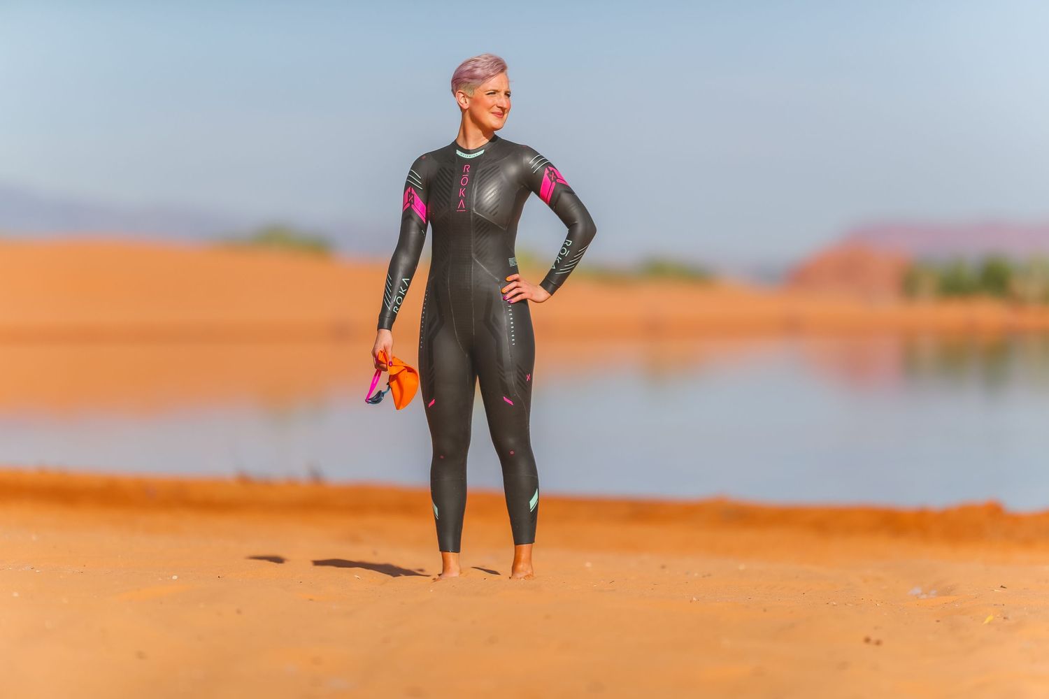 Female triathlete in wetsuit holding swim gear standing on sandy desert shoreline.