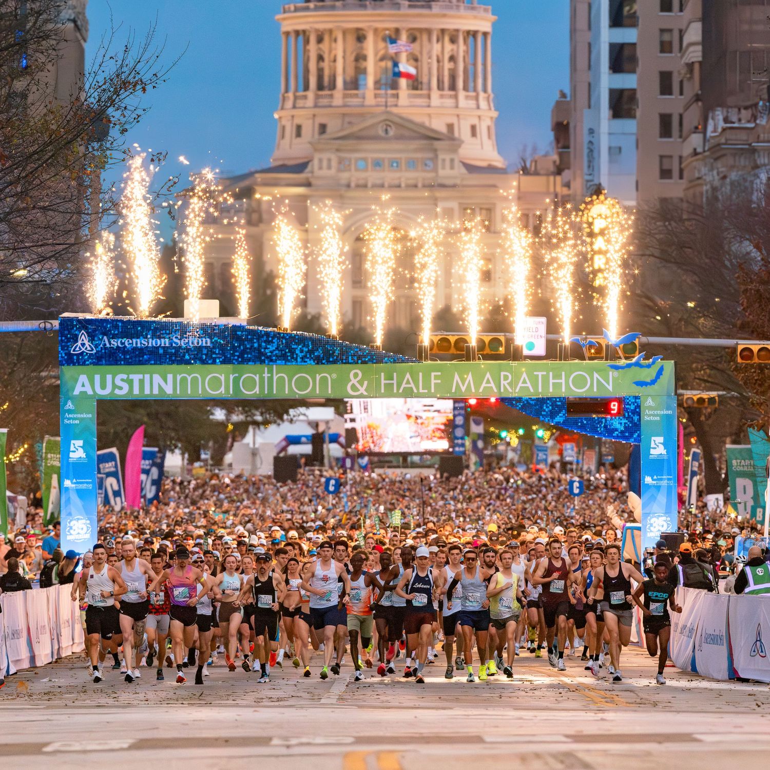 Massive crowd of runners at Austin Marathon start line with fireworks and illuminated capitol building.