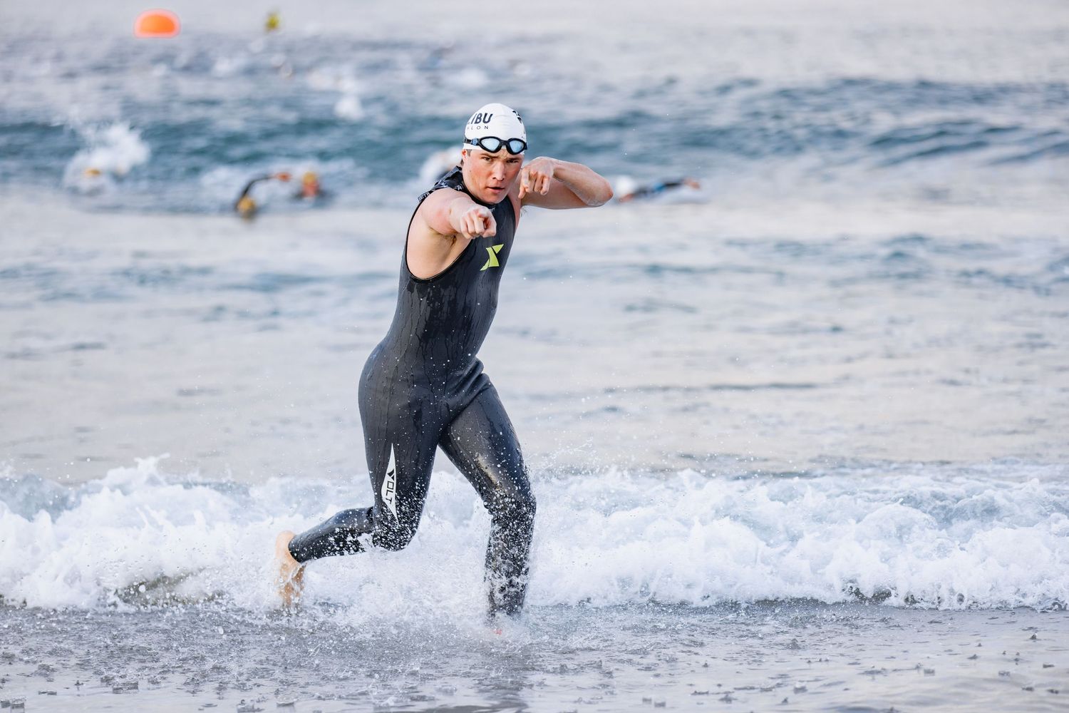 Triathlete in black wetsuit and goggles sprints out of ocean waves during a competitive swim event.