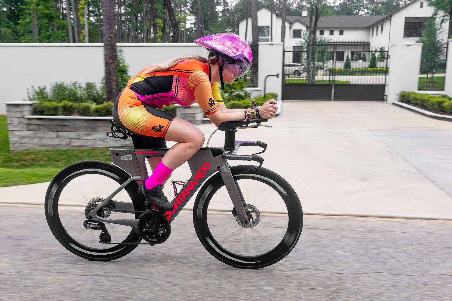 Female cyclist racing in bright pink and yellow kit on triathlon bike along tree-lined road.