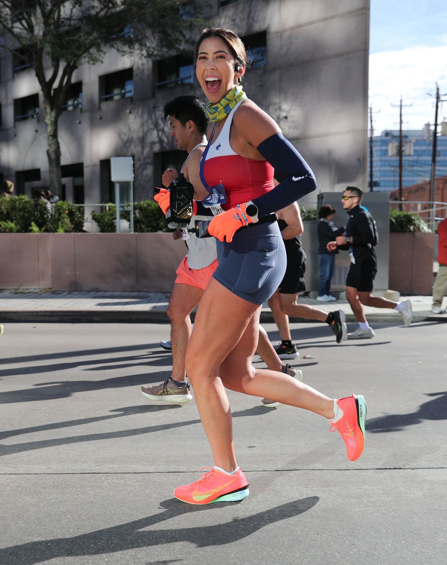 Smiling female runner in red top and orange Nike shoes races joyfully through city streets.