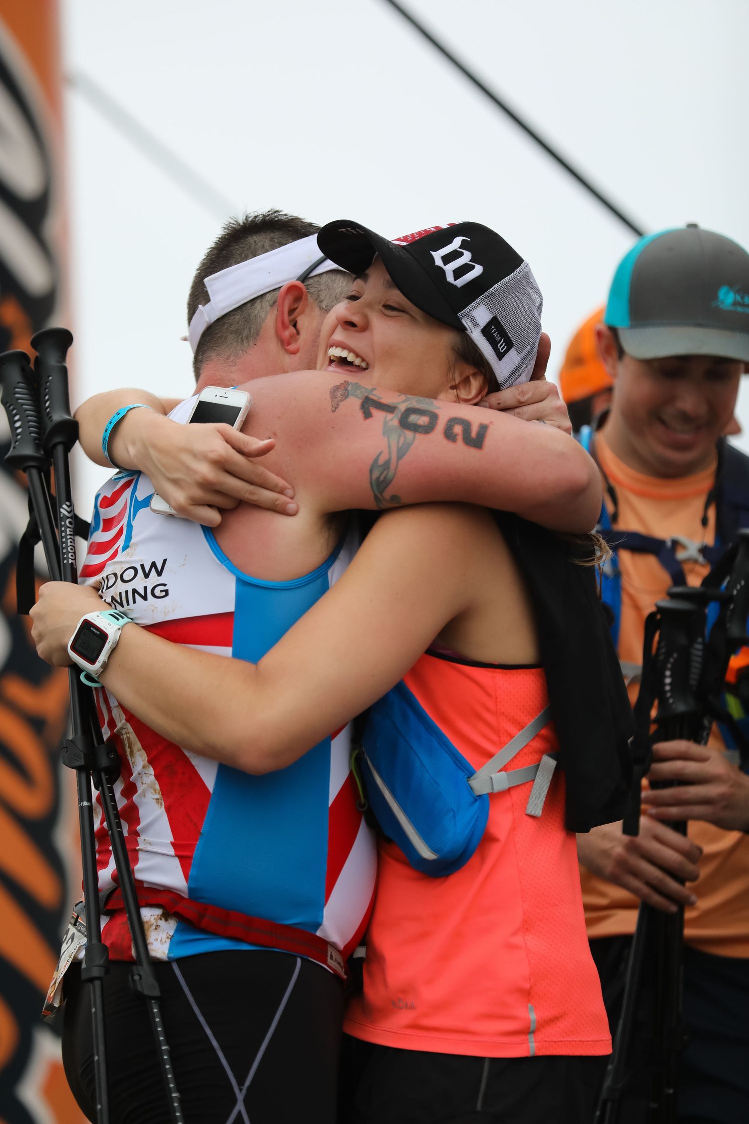 Two athletes share emotional hug at finish line of endurance race event outdoors.
