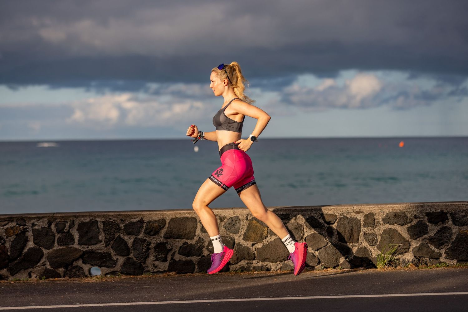 Female athlete running in pink shorts and sports bra along coastal road.