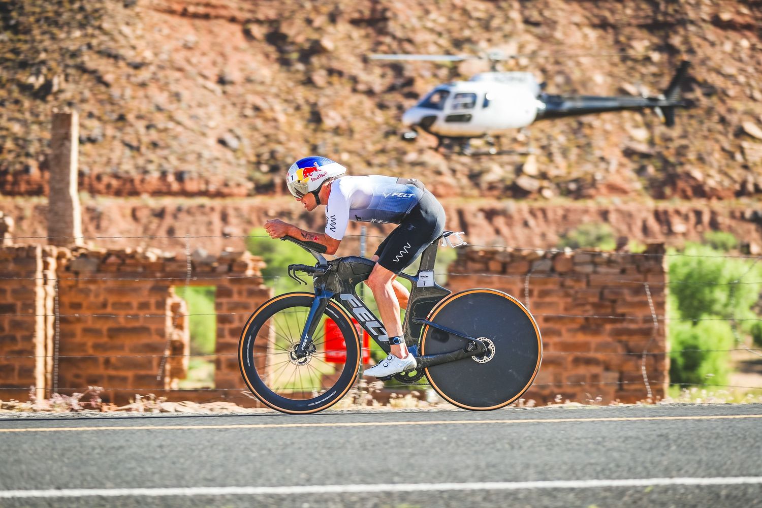 Cyclist in aerodynamic position races on road with helicopter flying overhead.