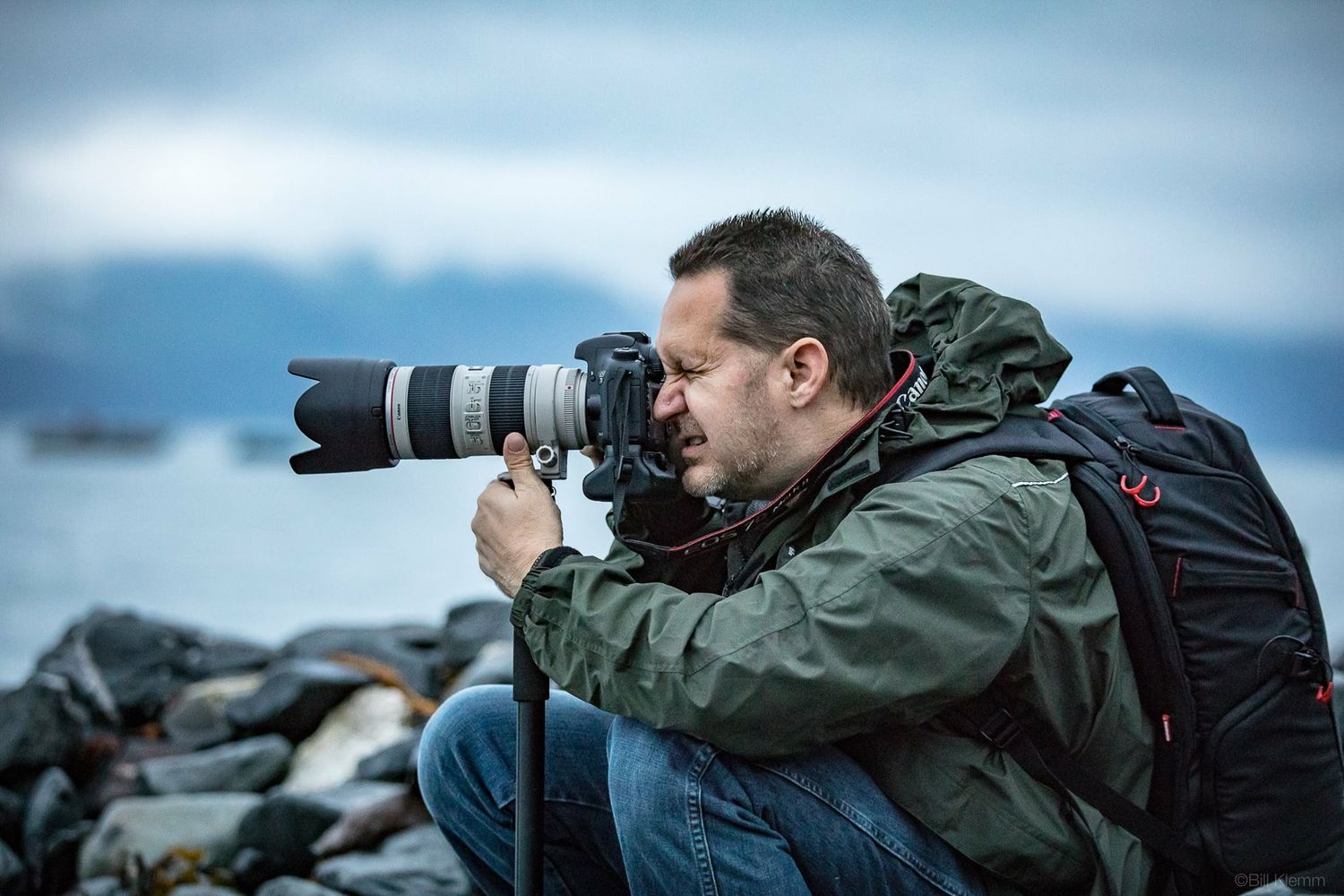 Male photographer shooting with telephoto lens on rocky coastal shoreline.