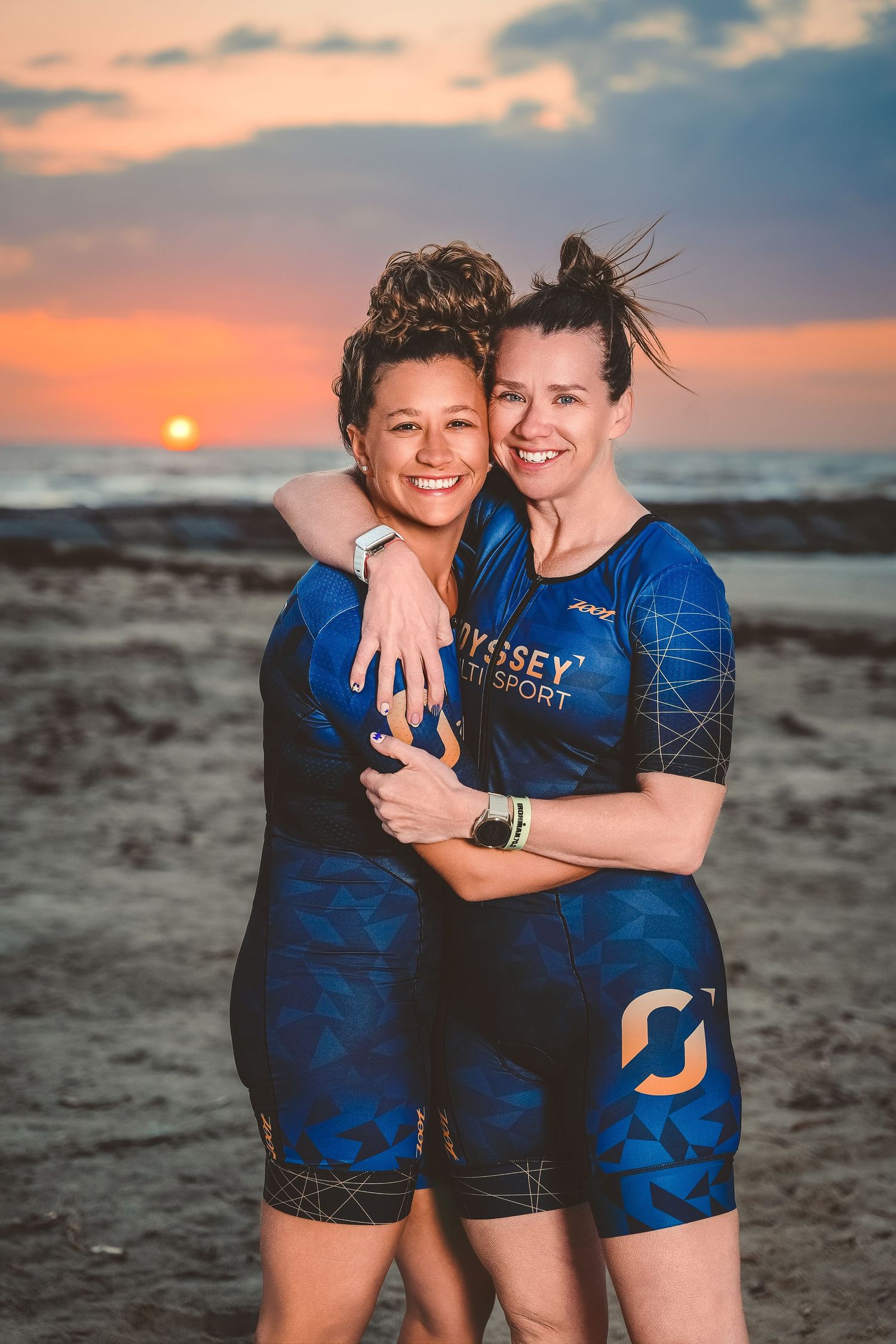 Two women in blue Odyssey triathlon suits hugging and smiling on beach during vibrant sunset.