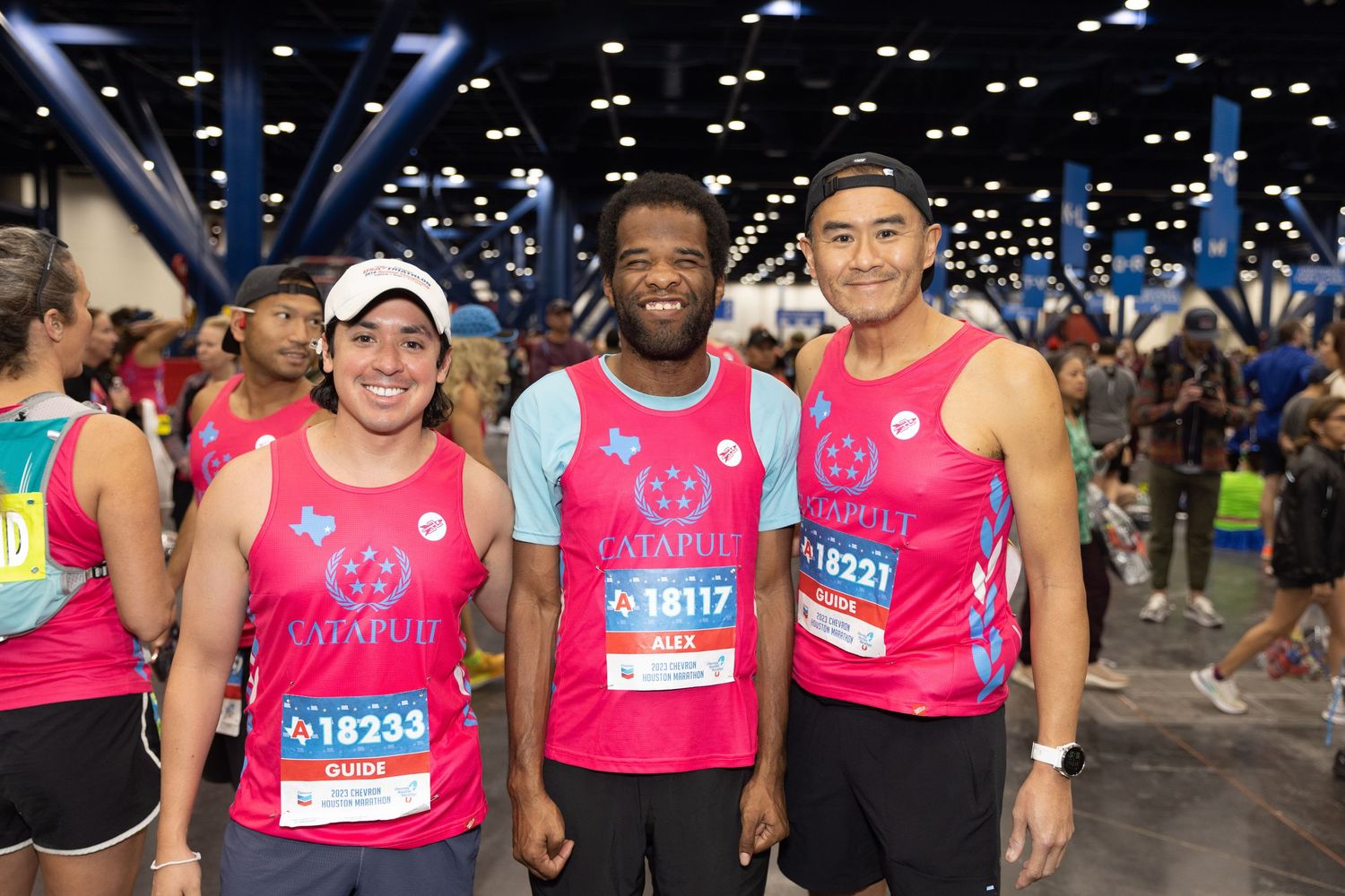 Three smiling runners in pink race jerseys posing together at marathon starting area.