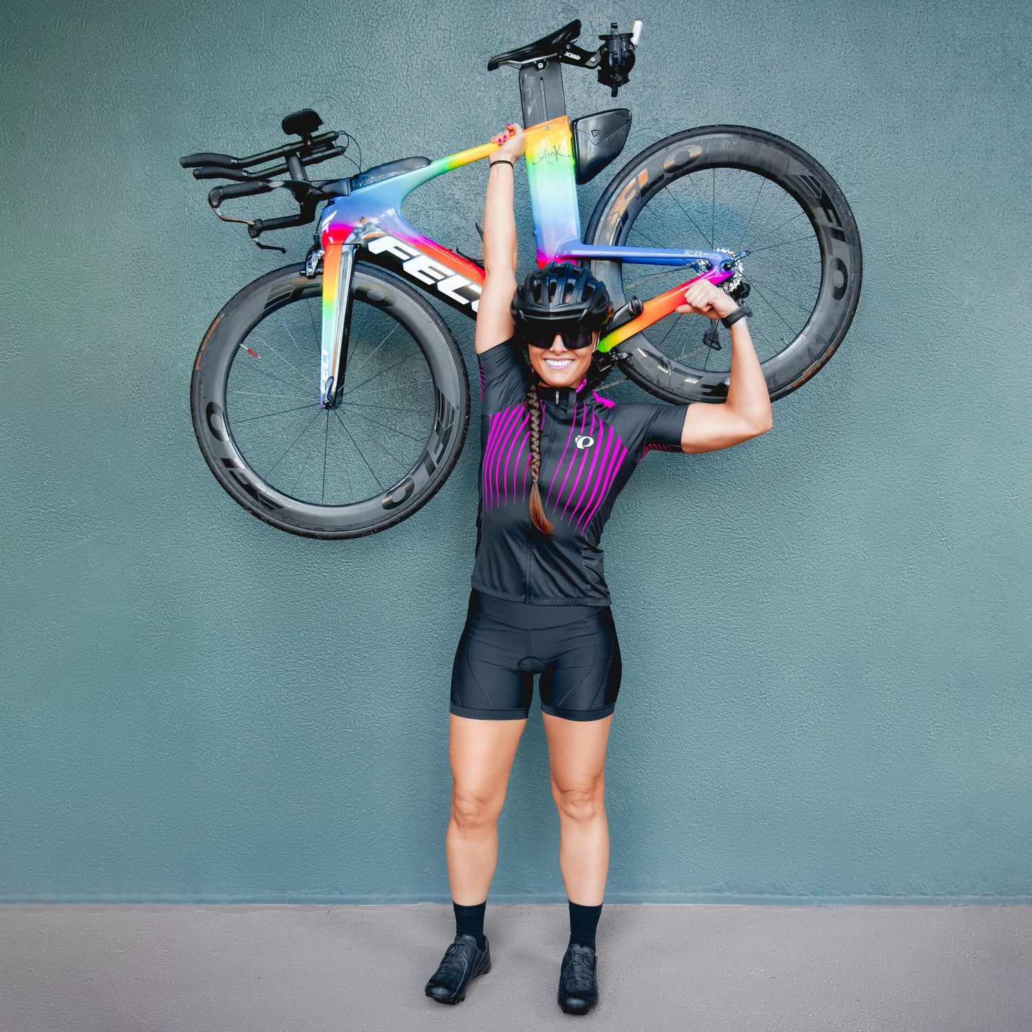 Woman cyclist lifting colorful Trek bike overhead wearing black cycling kit against gray wall.