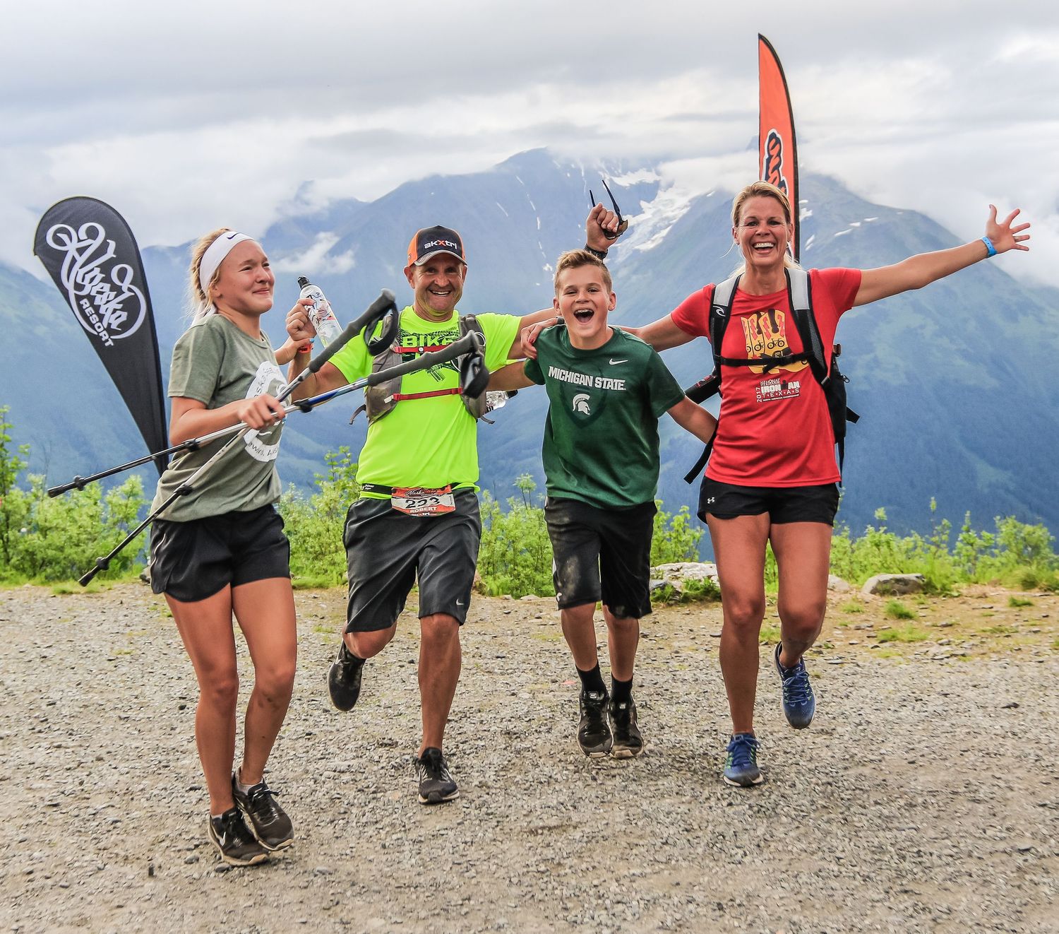 Four jubilant trail runners celebrate at mountain summit with flags and scenic alpine backdrop.