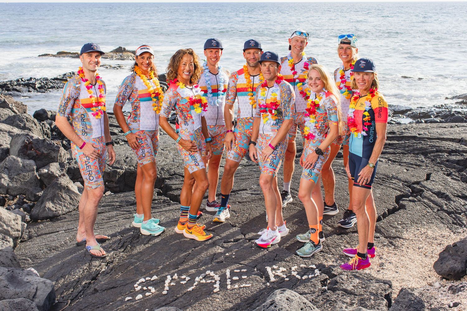 Group of athletes in colorful floral triathlon gear posing on volcanic rocks near the ocean.