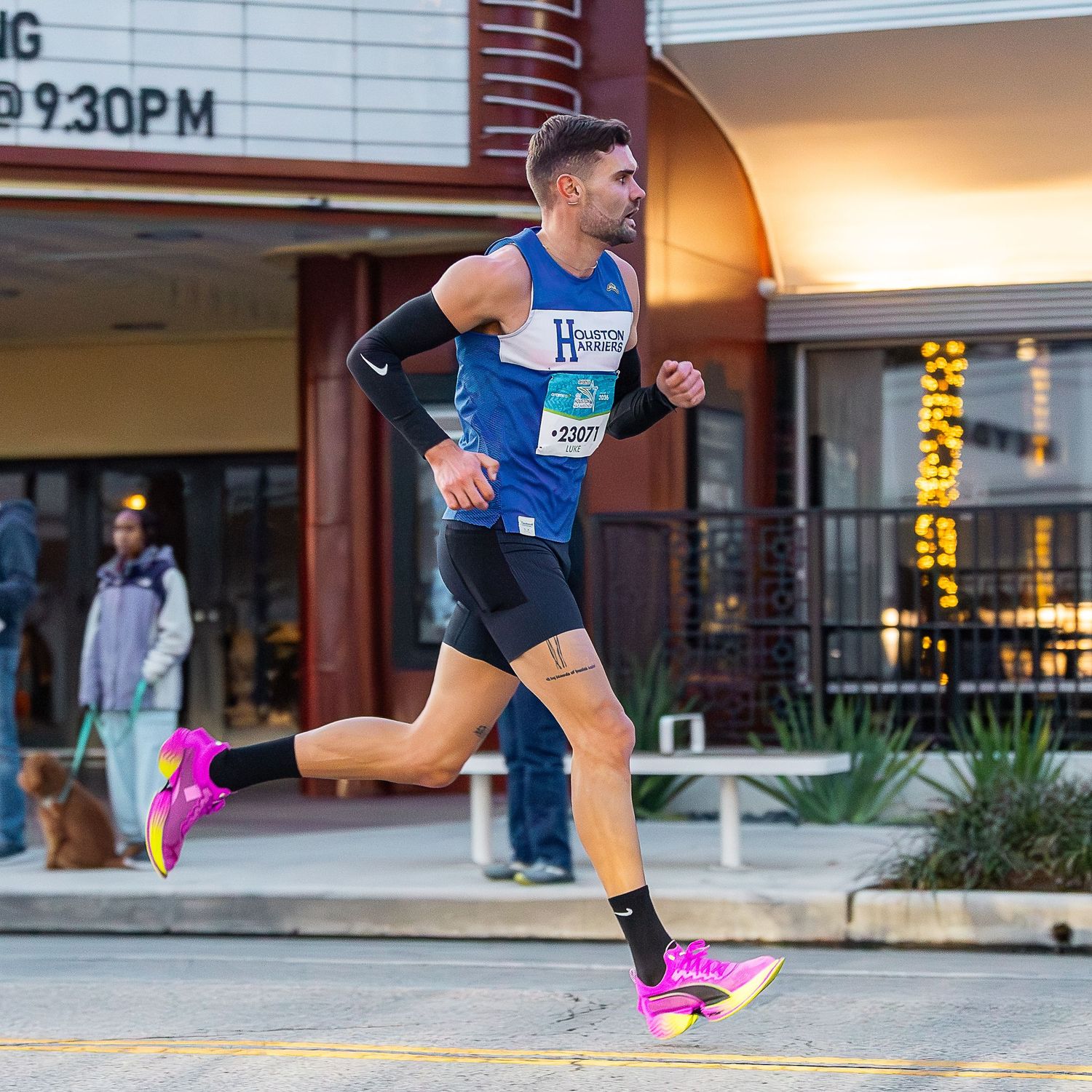 Male runner in blue vest sprints past a theater marquee wearing bright pink racing shoes.