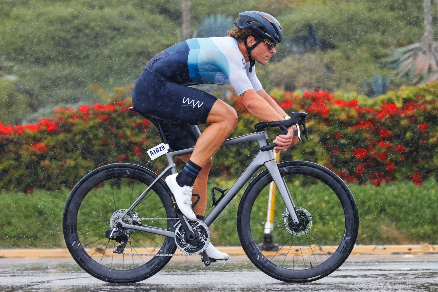 Cyclist in blue kit rides a gray road bike on wet pavement during a triathlon cycling segment.