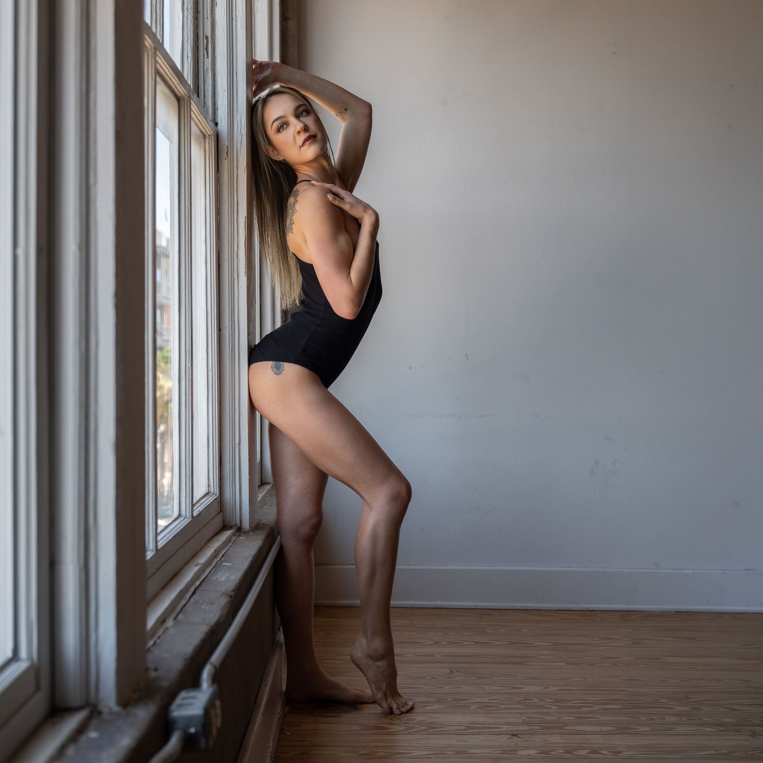Young woman in black bodysuit posing gracefully by large windows on wooden floor indoors.