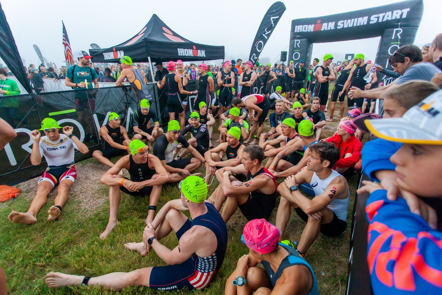 Large group of swimmers in green caps sitting at Ironman triathlon swim start area.