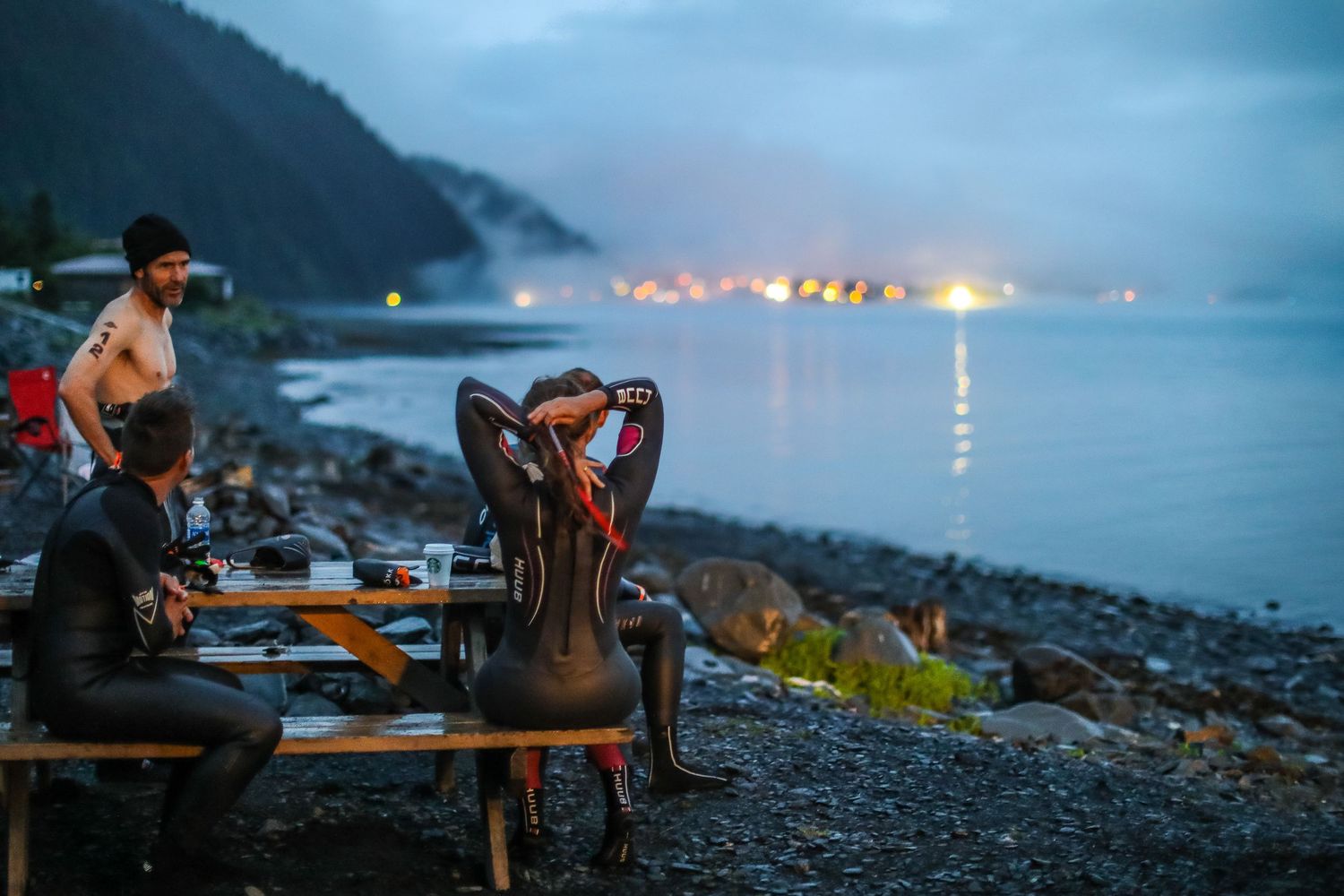 Triathletes in wetsuits prepare at a rocky shoreline transition area at dusk before race.