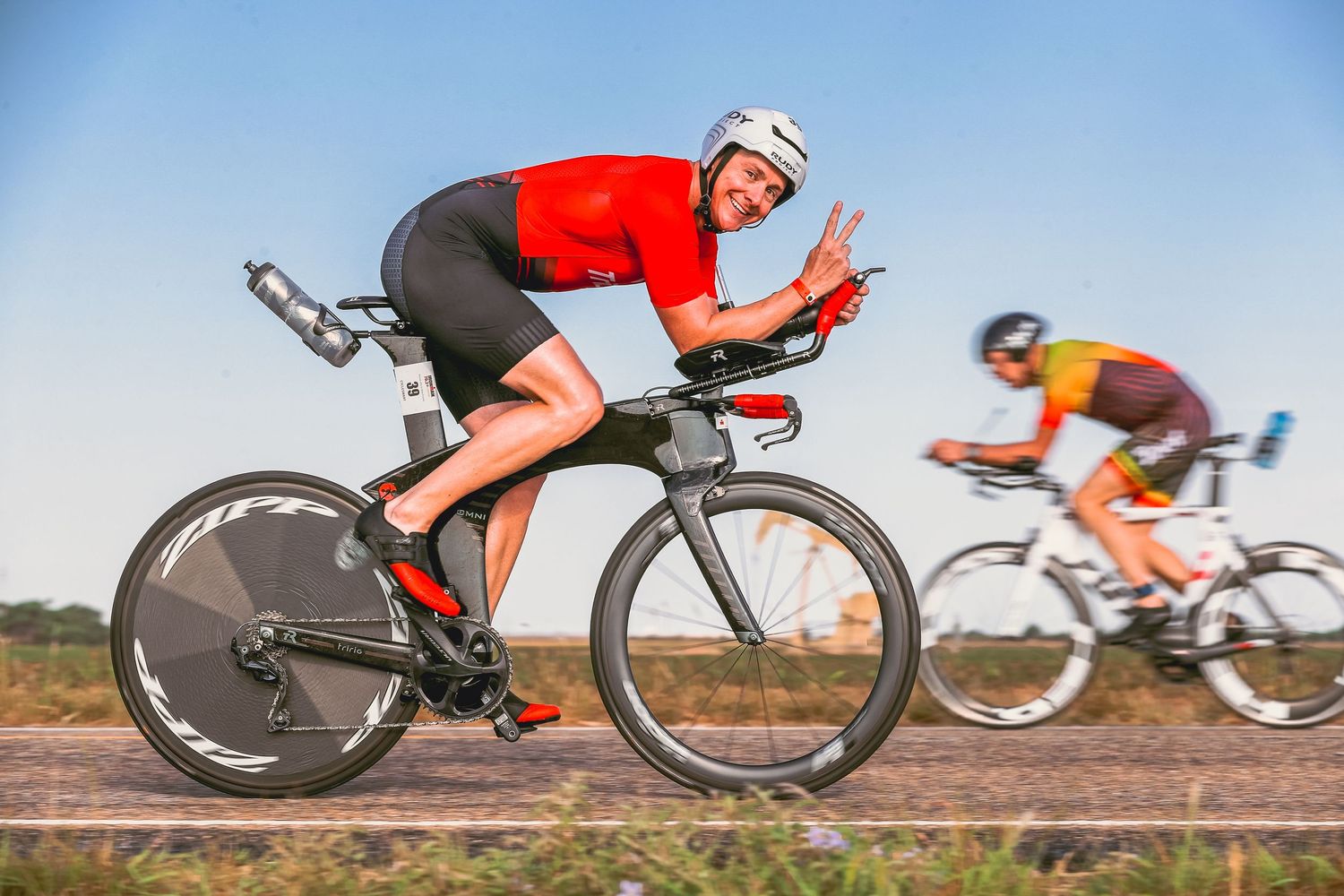 Smiling cyclist in red jersey gives peace sign while riding a time trial bike in a triathlon race.