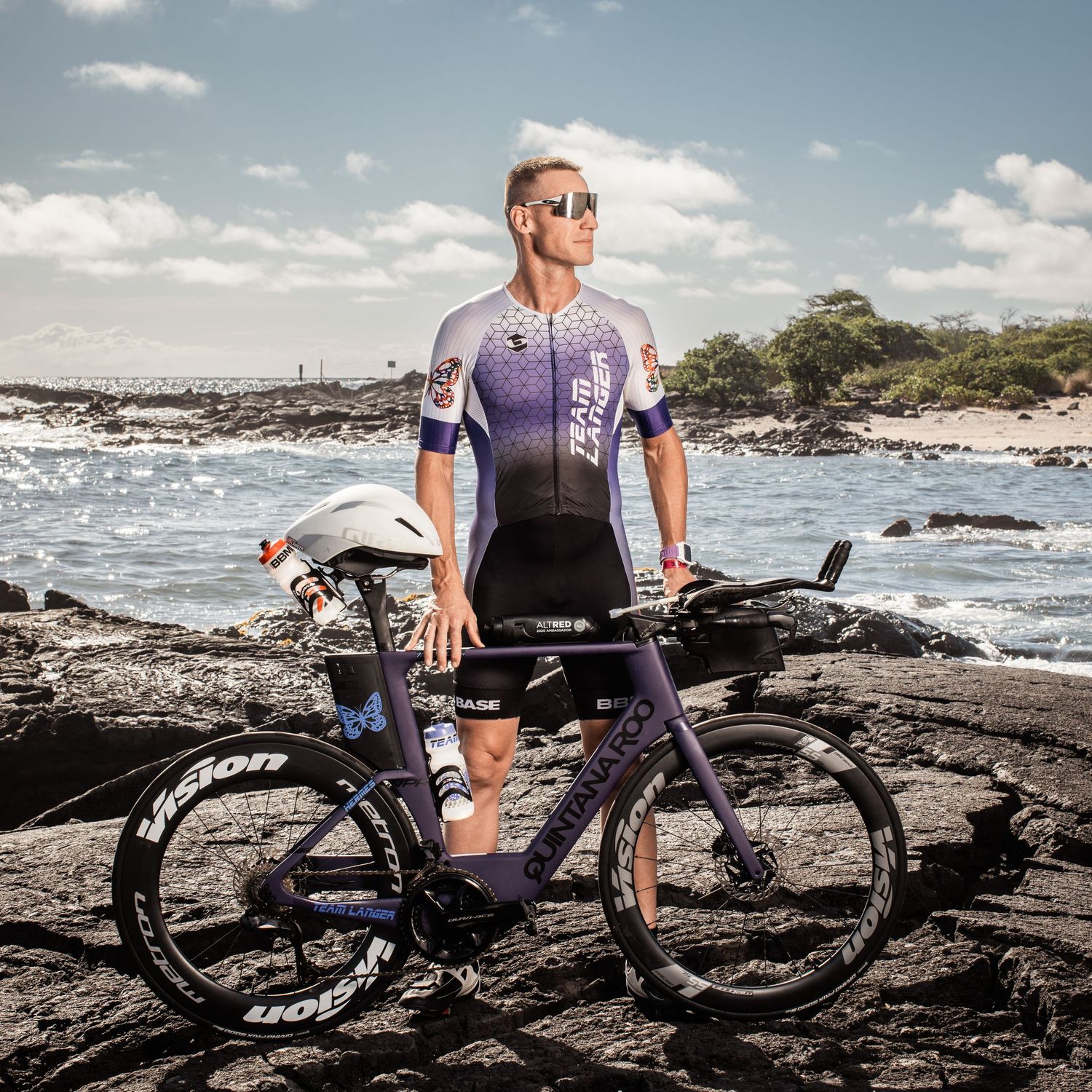 Male triathlete in purple cycling kit standing with bike on rocky Hawaiian coastline.