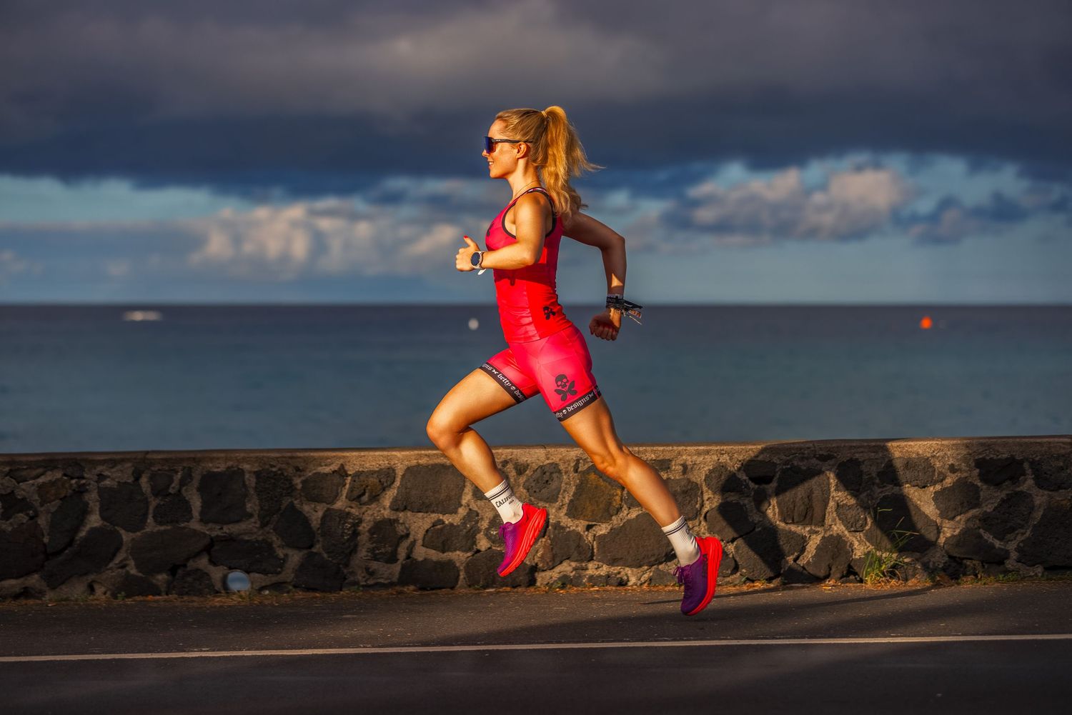 Female triathlete in red running suit sprinting along coastal road with ocean and dramatic sky backdrop.