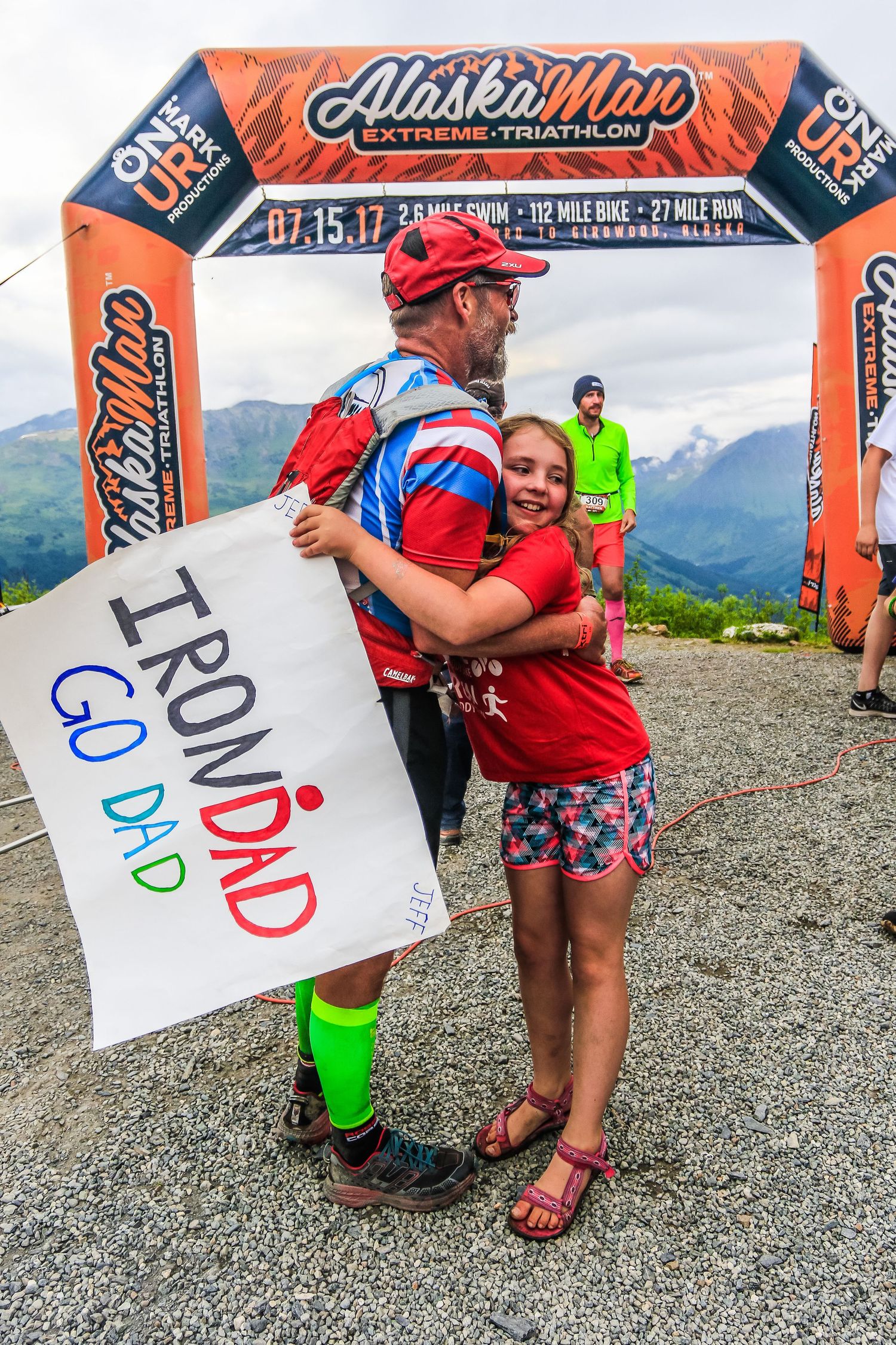 Emotional triathlete hugs young daughter holding IronDad sign at AlaskaMan Extreme Triathlon finish.