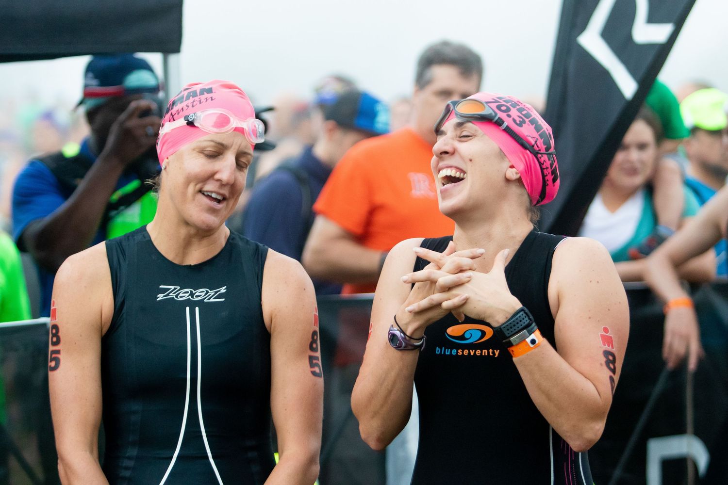 Two female triathletes in wetsuits and pink swim caps laugh together before race start.
