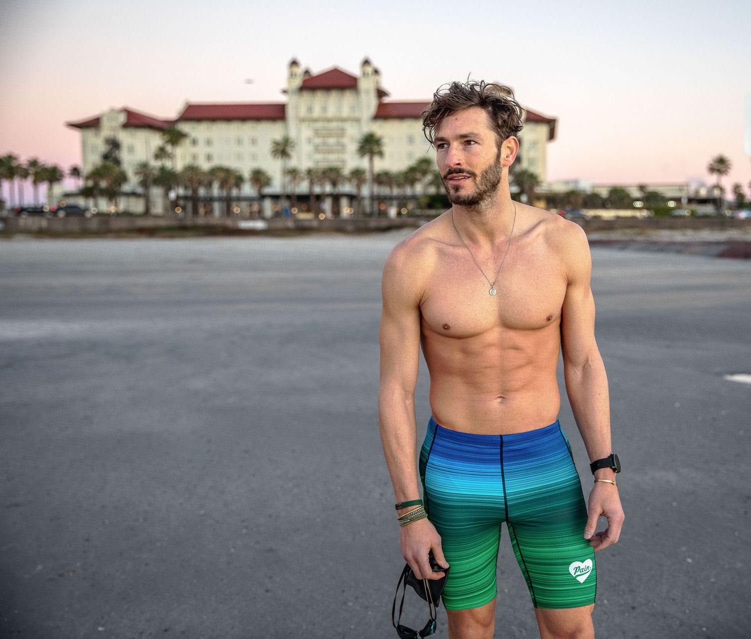 Fit shirtless man in colorful swim shorts standing outdoors near a historic beachfront building.