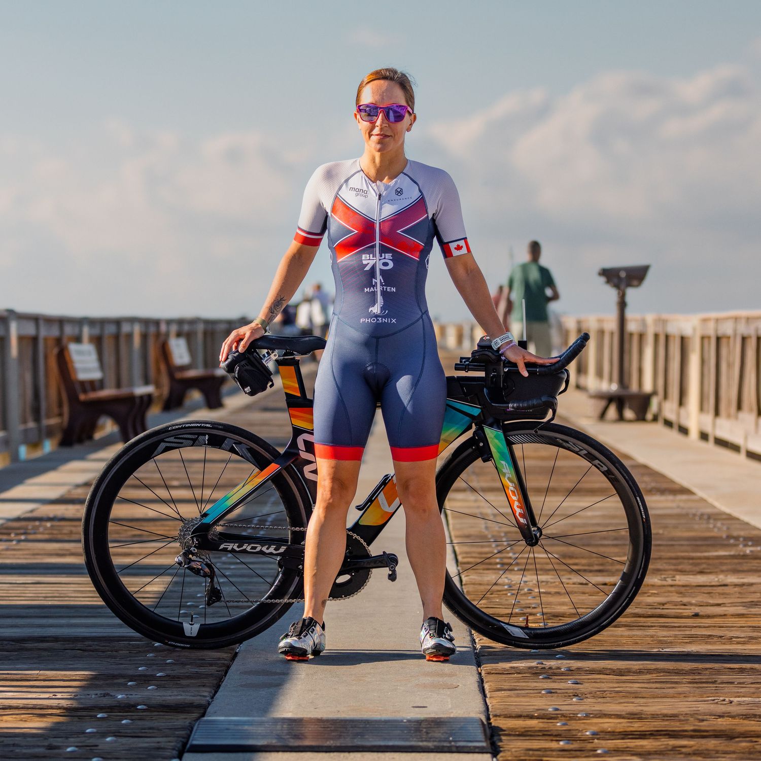 Female triathlete in British kit standing with a triathlon bike on a sunny wooden pier.