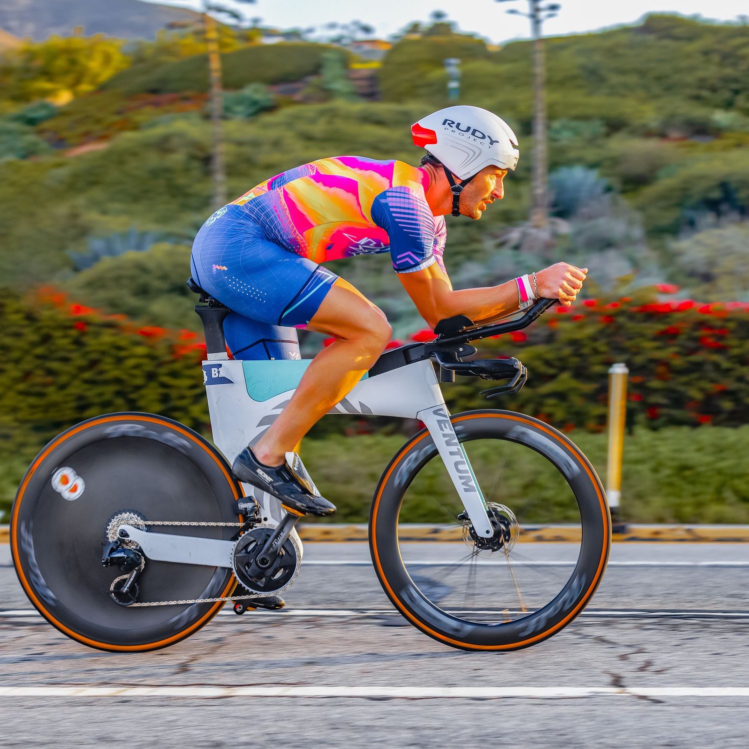 Cyclist wearing colorful pink and orange jersey riding a time trial bike with disc wheel on a road course.