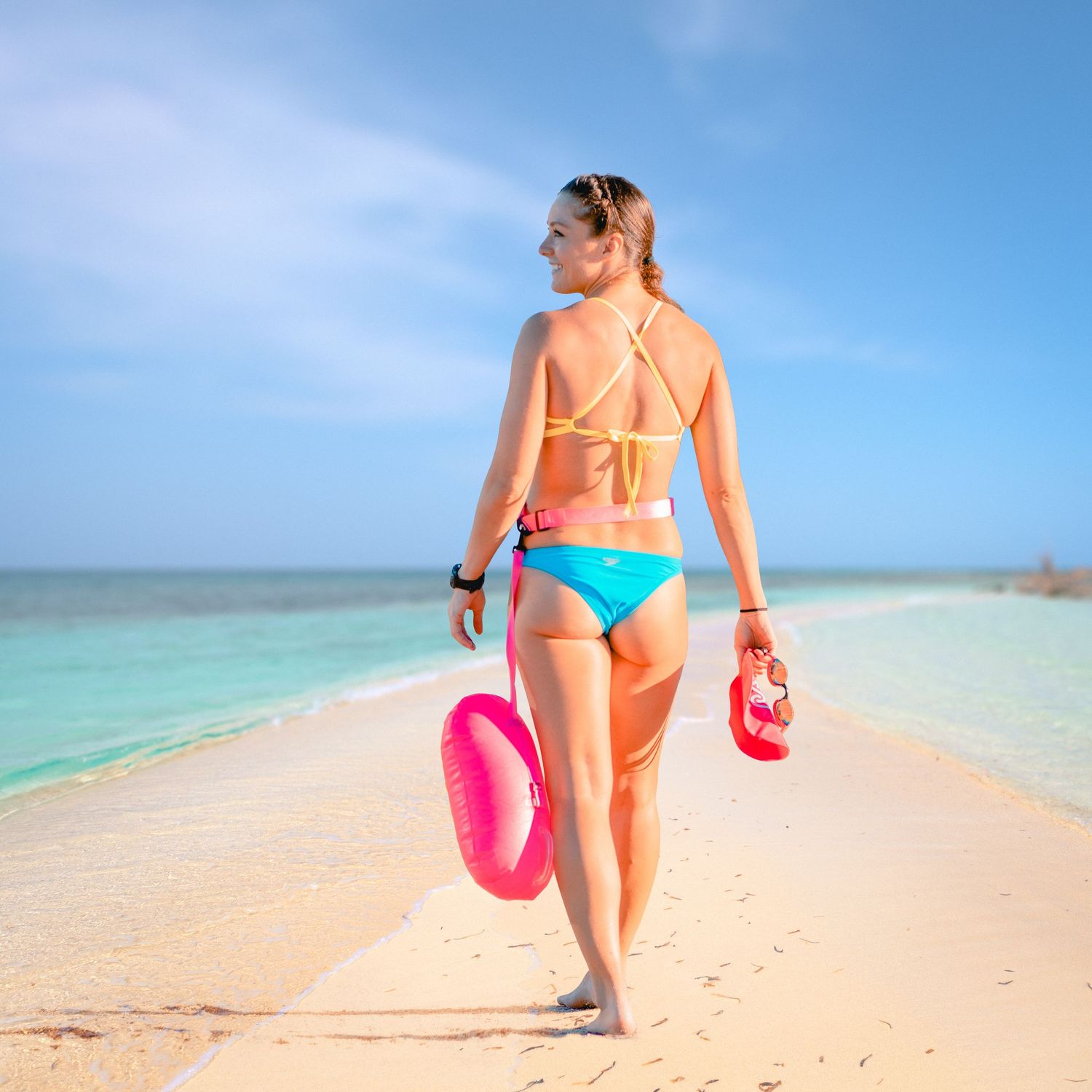Woman in colorful bikini walking on tropical beach carrying pink float ring and red fins.