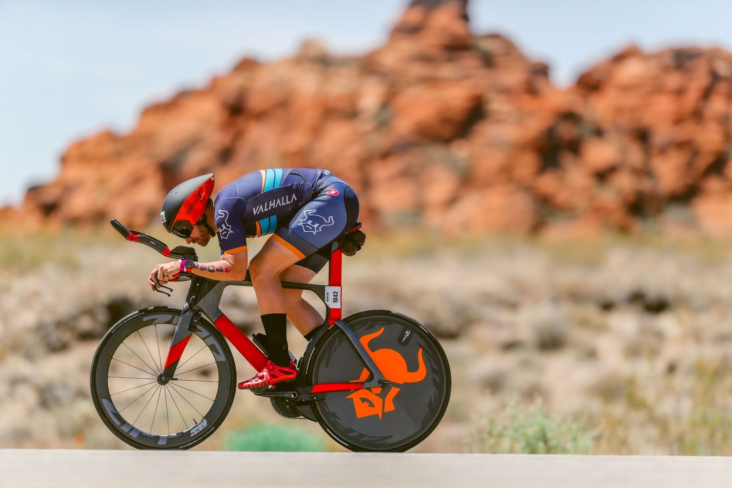 Triathlete in blue kit races on a red time trial bike past dramatic red rock desert scenery.