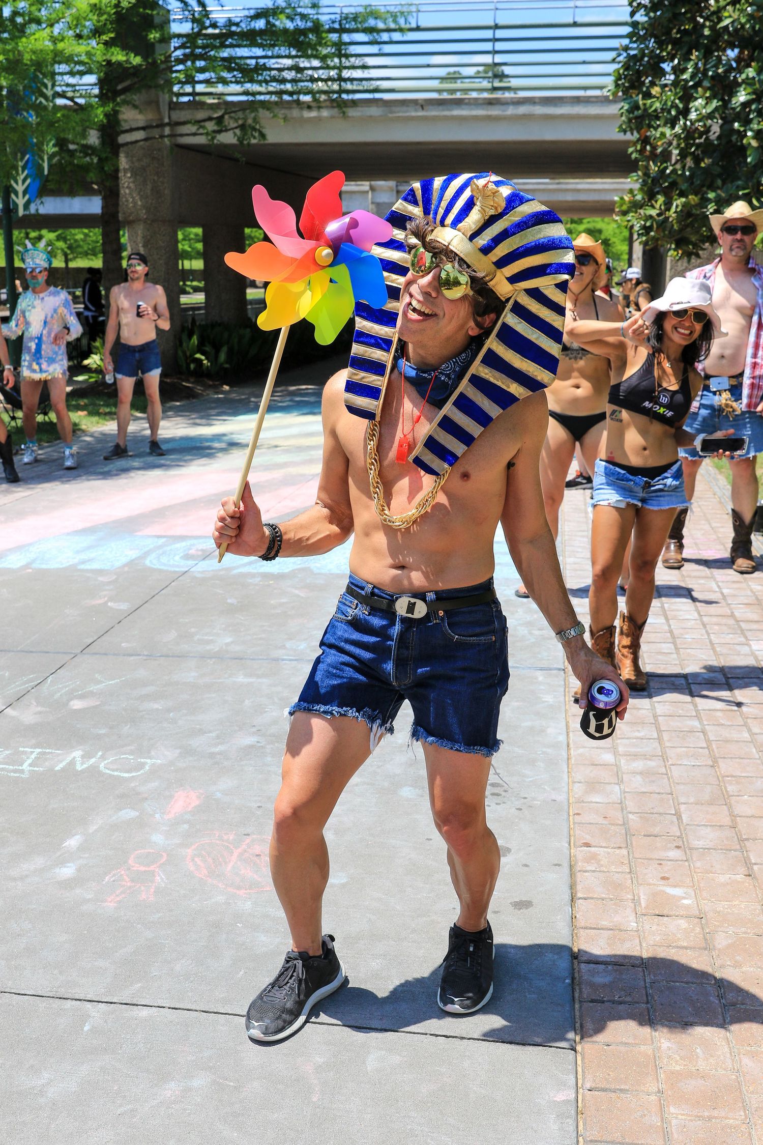 Shirtless man wearing Egyptian pharaoh costume holds colorful pinwheel at outdoor festival event.