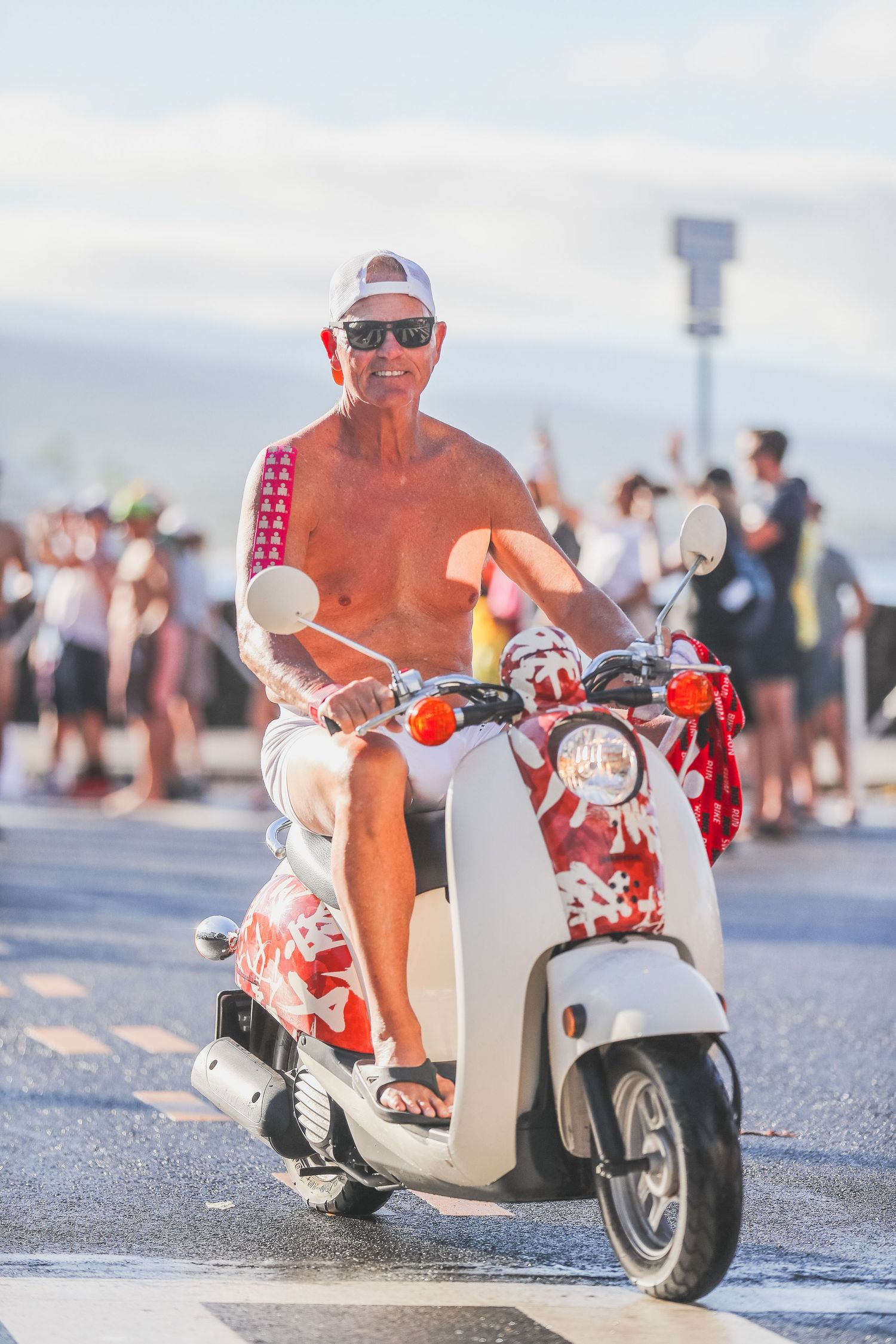 Shirtless Mike Reilly riding a decorated white scooter along a sunny race course road at the Kona Underpants Run.