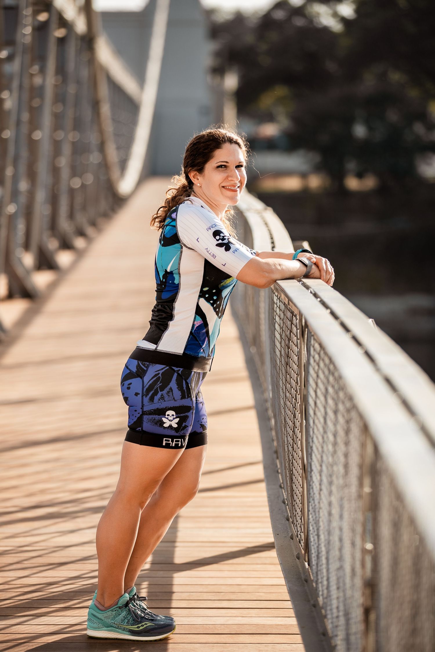 Female triathlete in blue skull cycling kit leaning on bridge railing smiling outdoors.