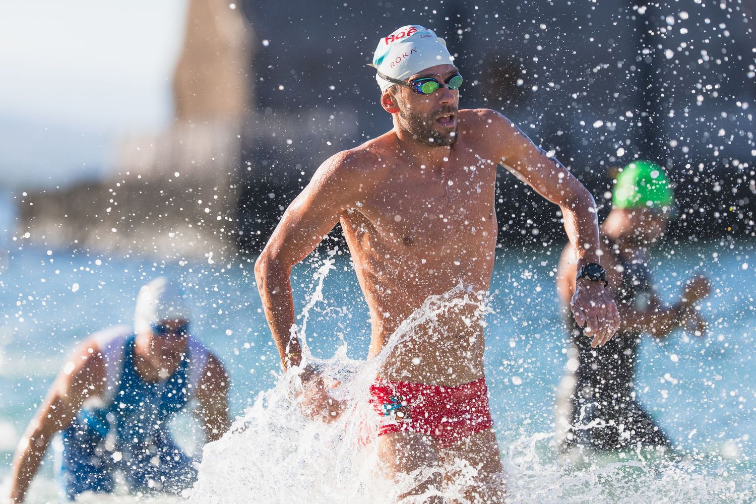 Male triathlete emerging from ocean waves wearing red swim trunks and goggles during an open water swim race.