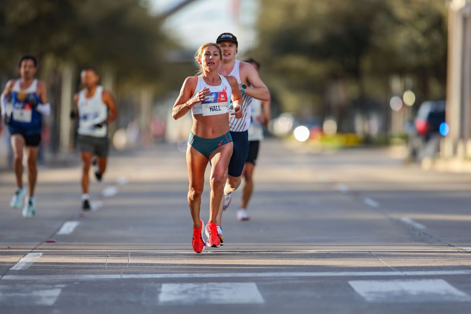 Female runner in white kit leads a group of athletes along a wide open road race course.