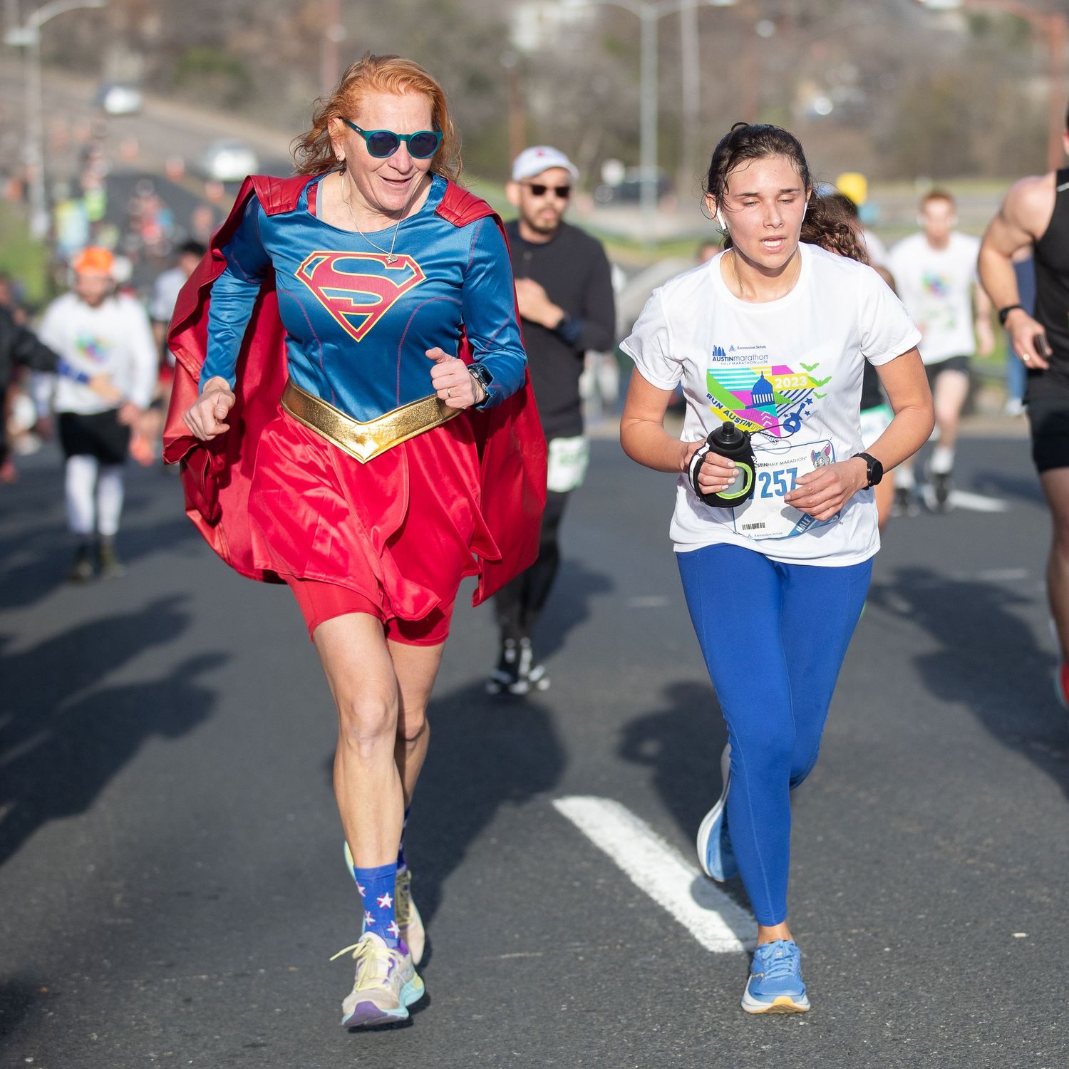 Woman in Supergirl costume runs alongside another female runner wearing bib 257 in a road race.