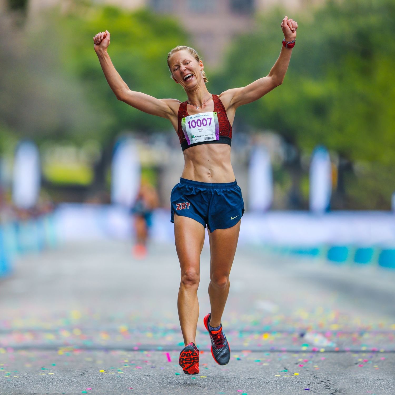 Female marathon runner celebrating victory with arms raised at finish line with bib number 10007.