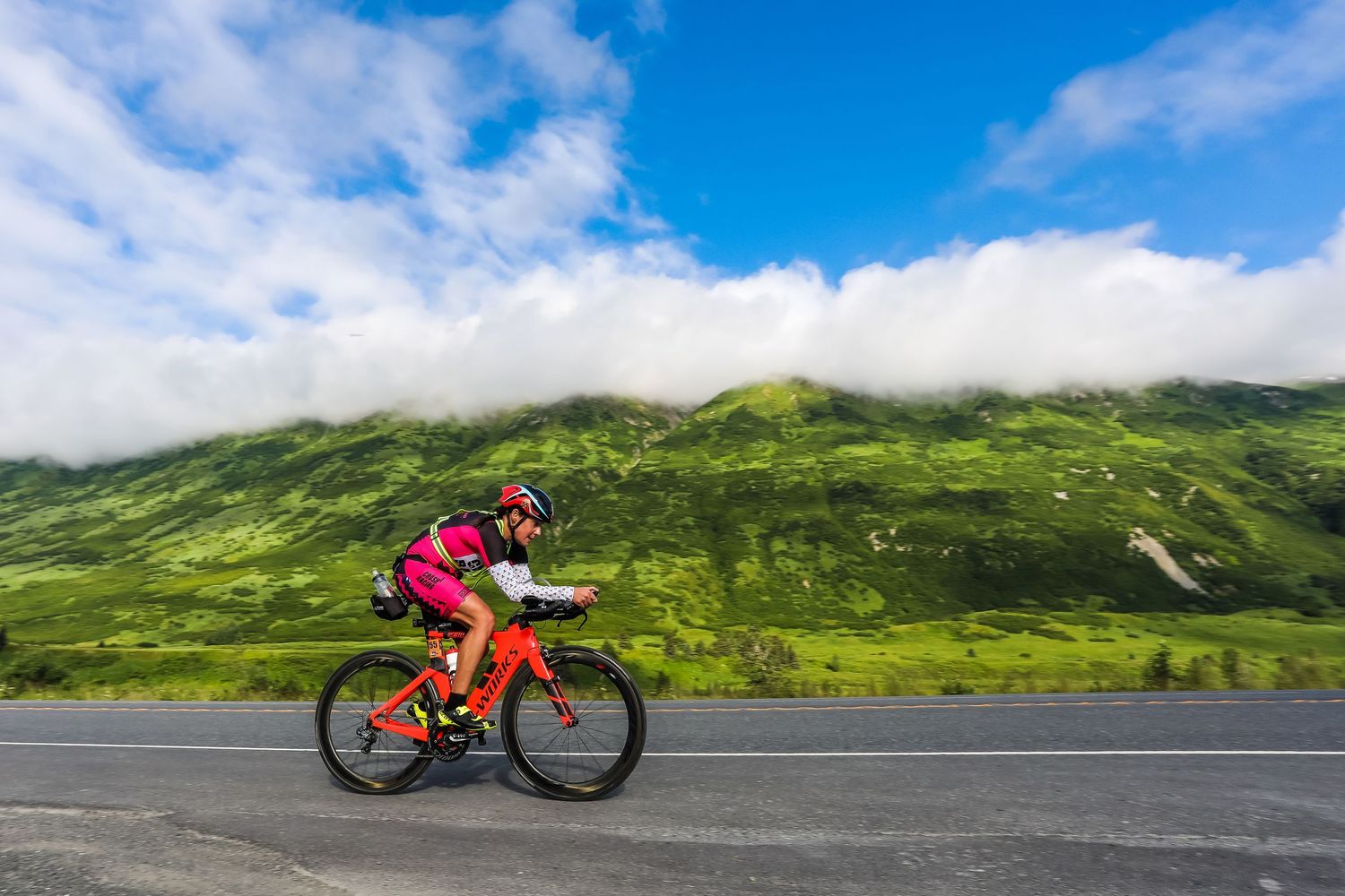 Female cyclist in pink kit rides a red road bike along a scenic mountain highway under blue skies at the inaugural Alaskaman Extreme Tri.