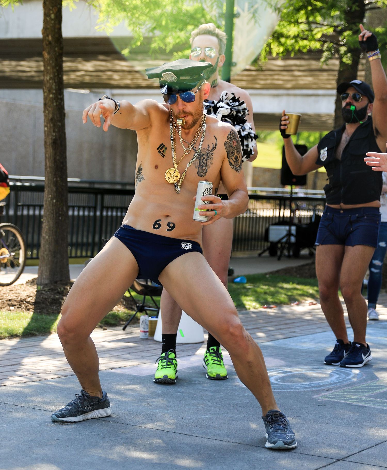 Shirtless tattooed man in briefs dances energetically while cheering at outdoor race.