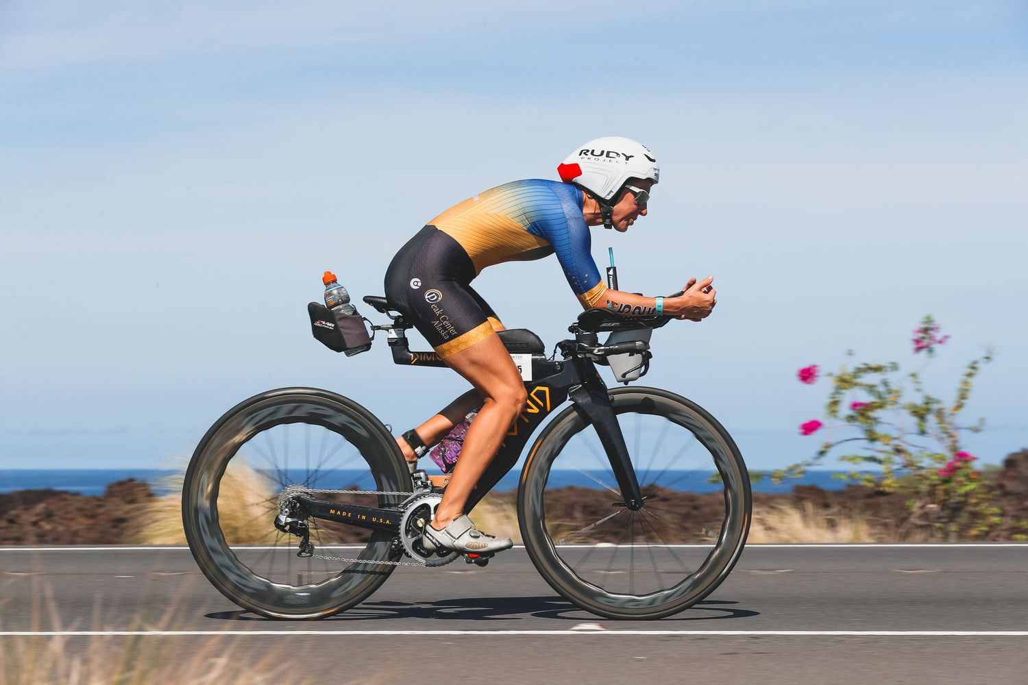 Cyclist in aerodynamic position riding a time trial bike along a coastal road during a triathlon race.