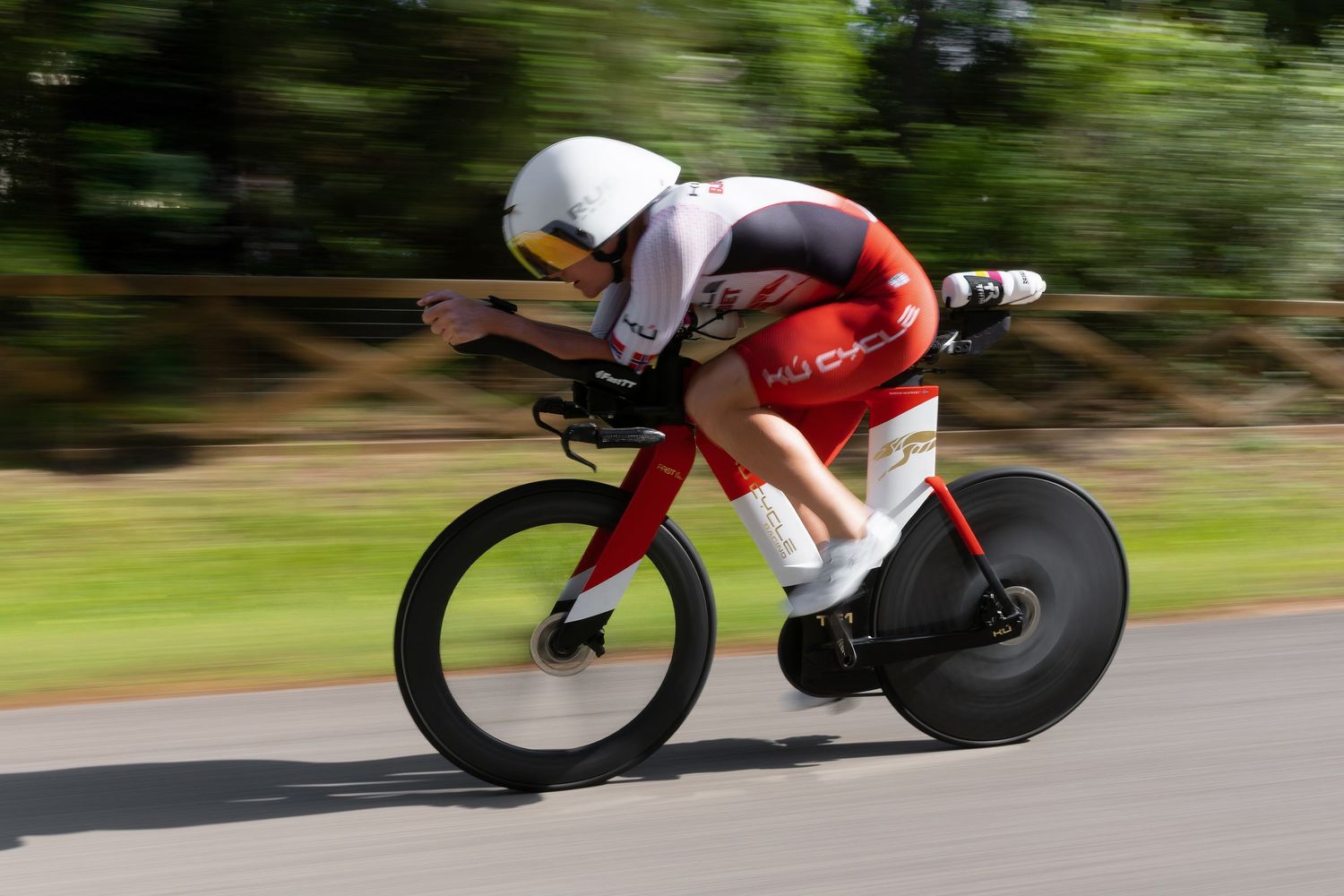 Cyclist in red and white kit riding aerodynamically on time trial bike with blurred green background.