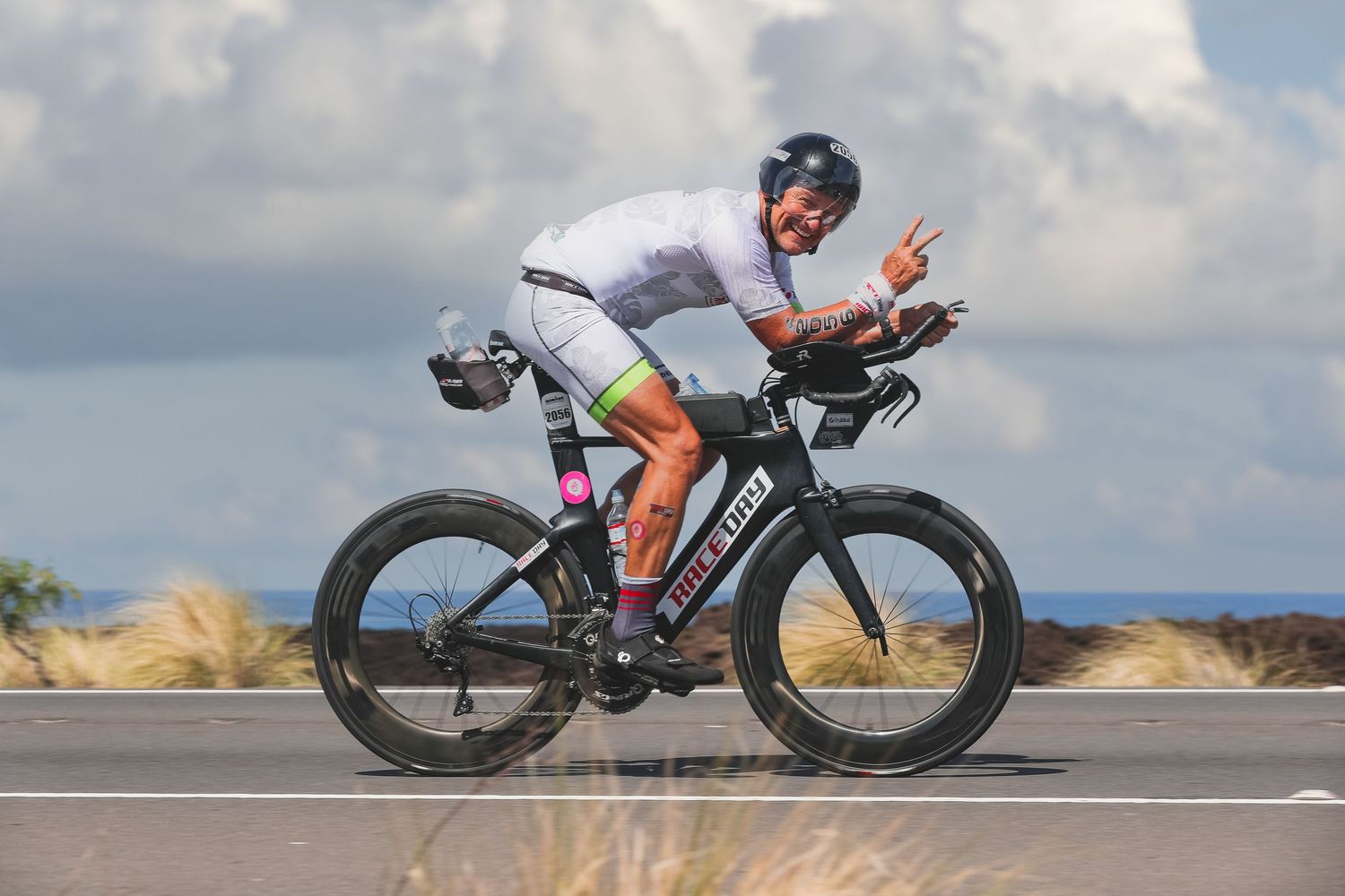 Male triathlete cycling in aero position giving peace sign on open road.