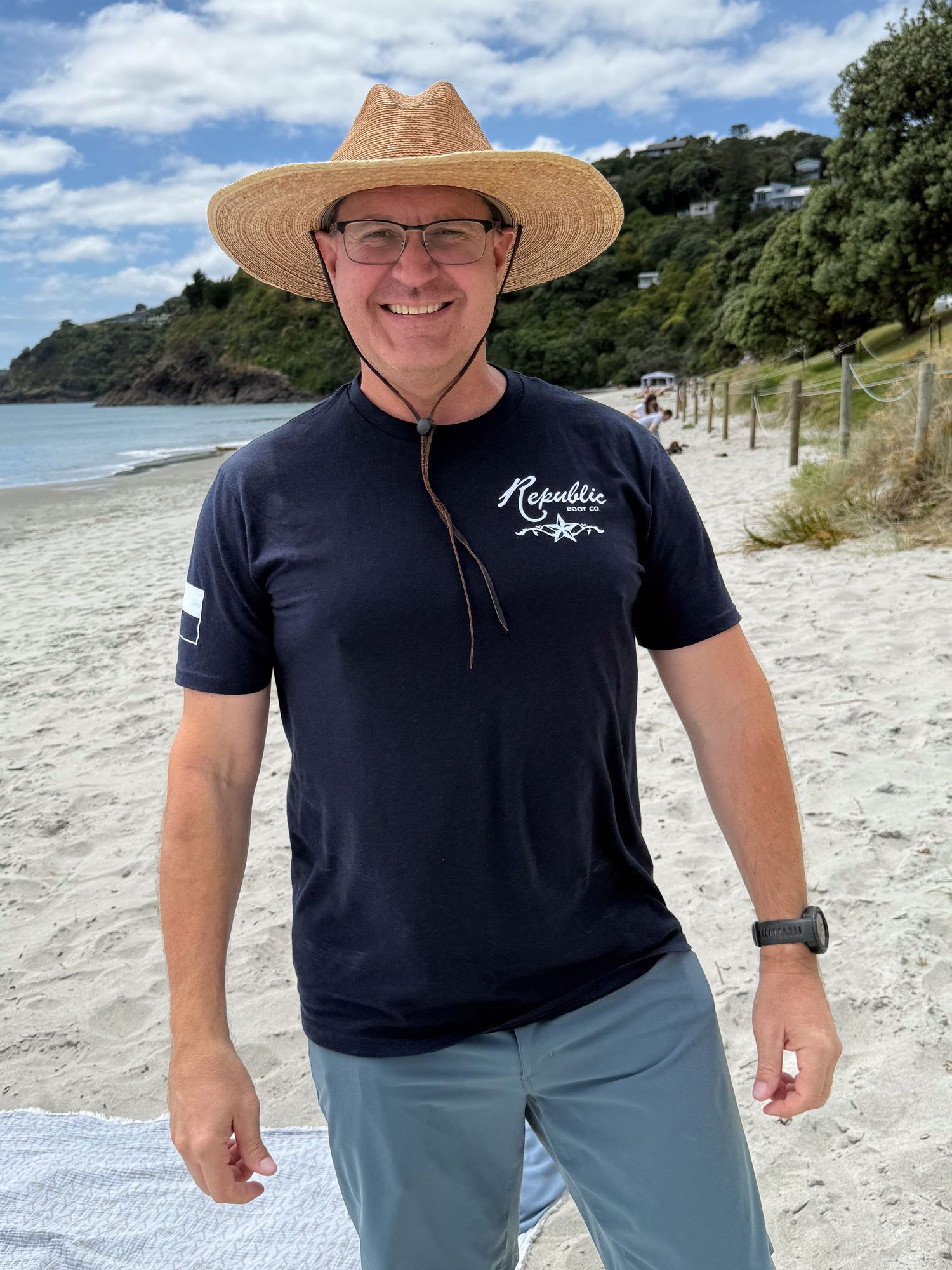 Man smiling on beach wearing straw hat and navy Republic shirt with gray shorts on sunny day.