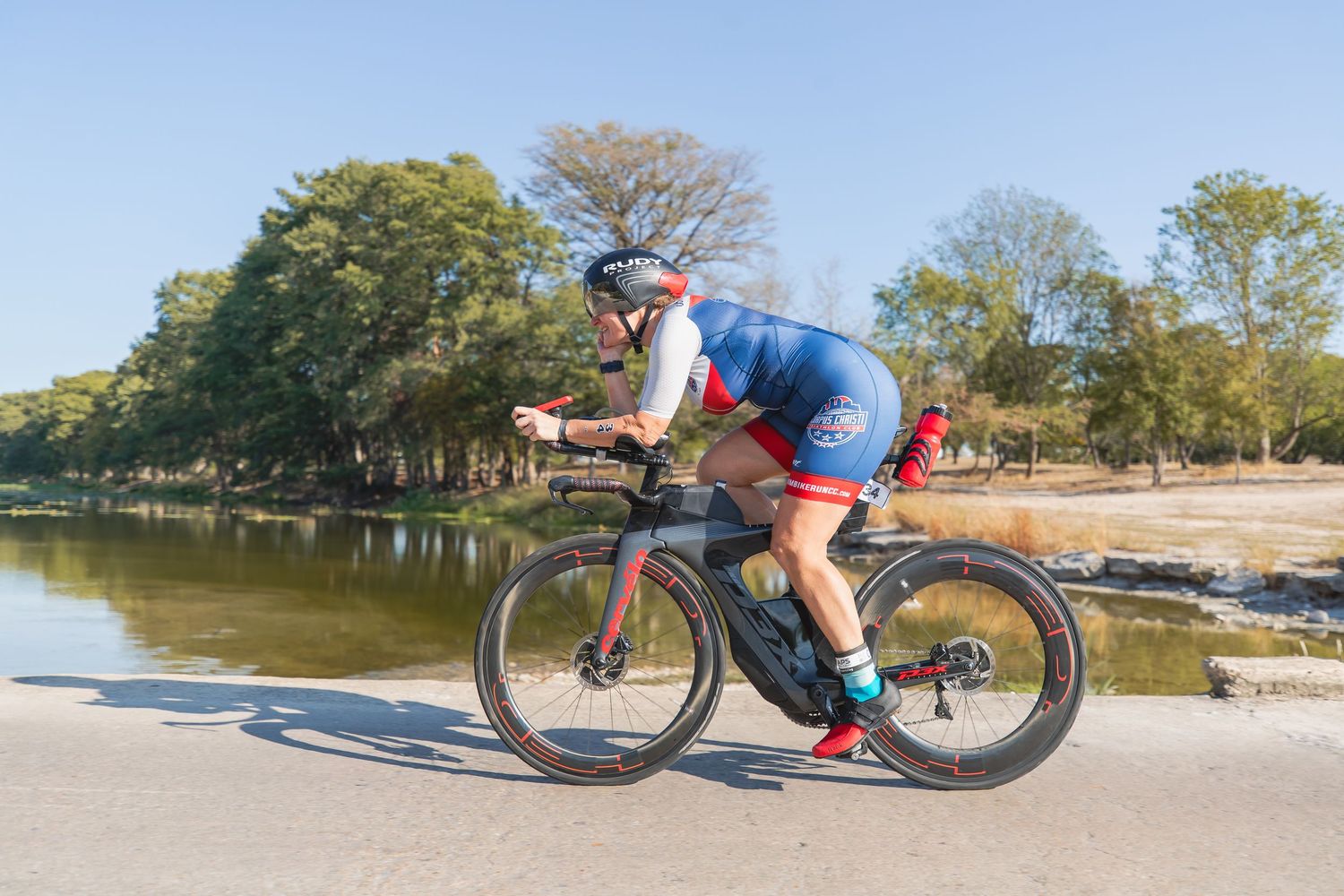 Cyclist in blue triathlon kit rides aerodynamically along a scenic riverside road during a triathlon race.