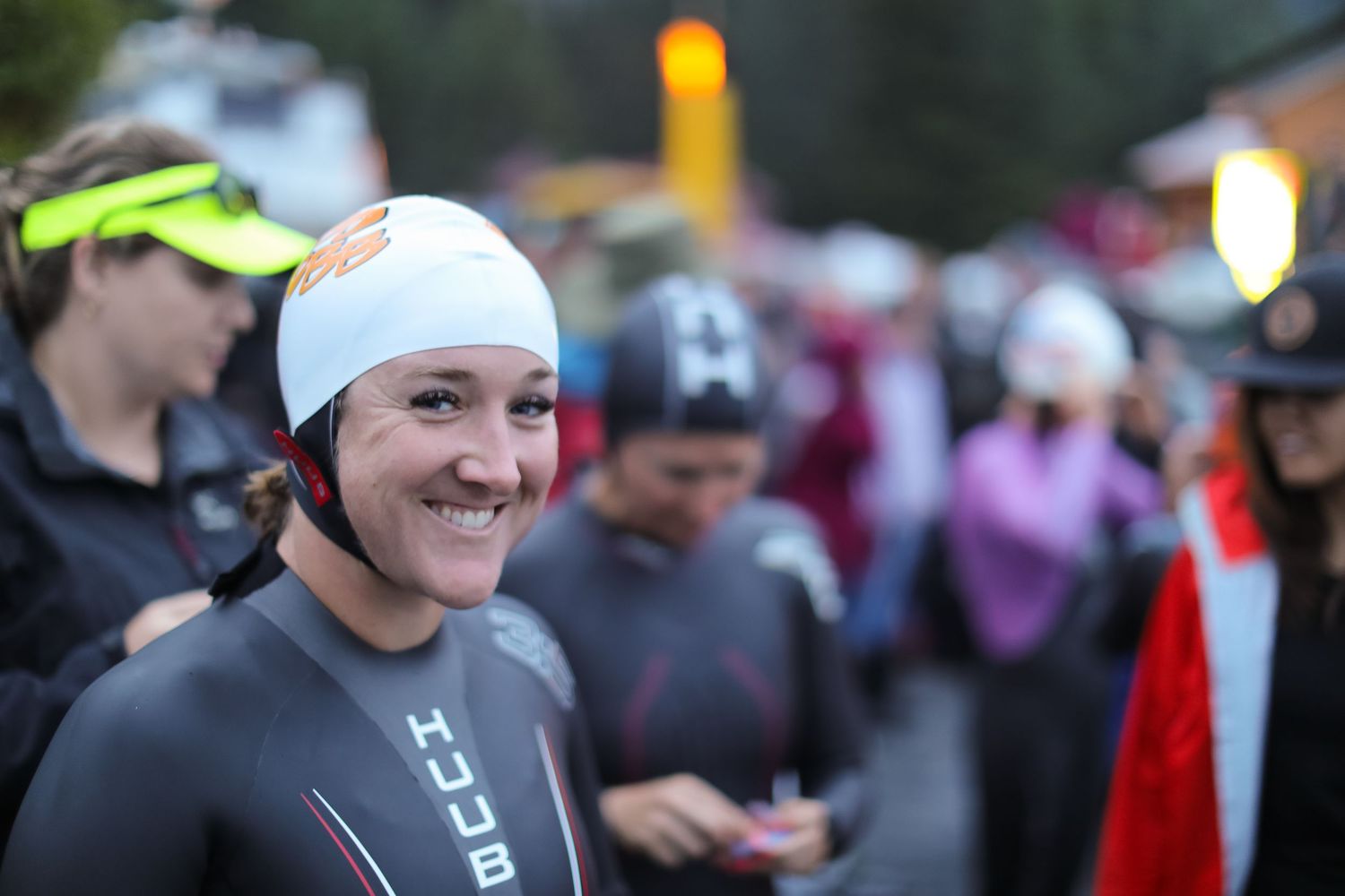 Smiling female triathlete in wetsuit and white swim cap at crowded race starting area.